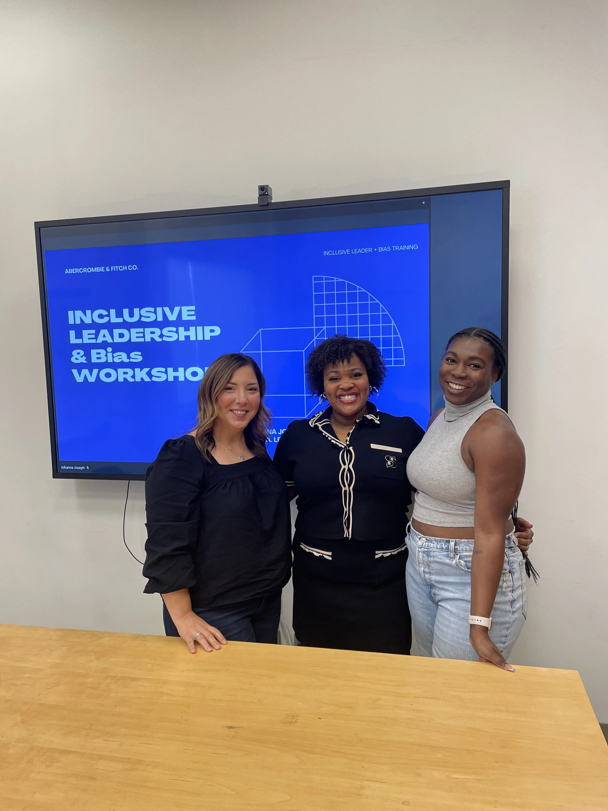Three women standing in front of a large digital screen that reads 'Inclusive Leadership & Bias Workshop' and shows a geometric graphic. The women are smiling and posing for the photo, with a plain white wall behind them.