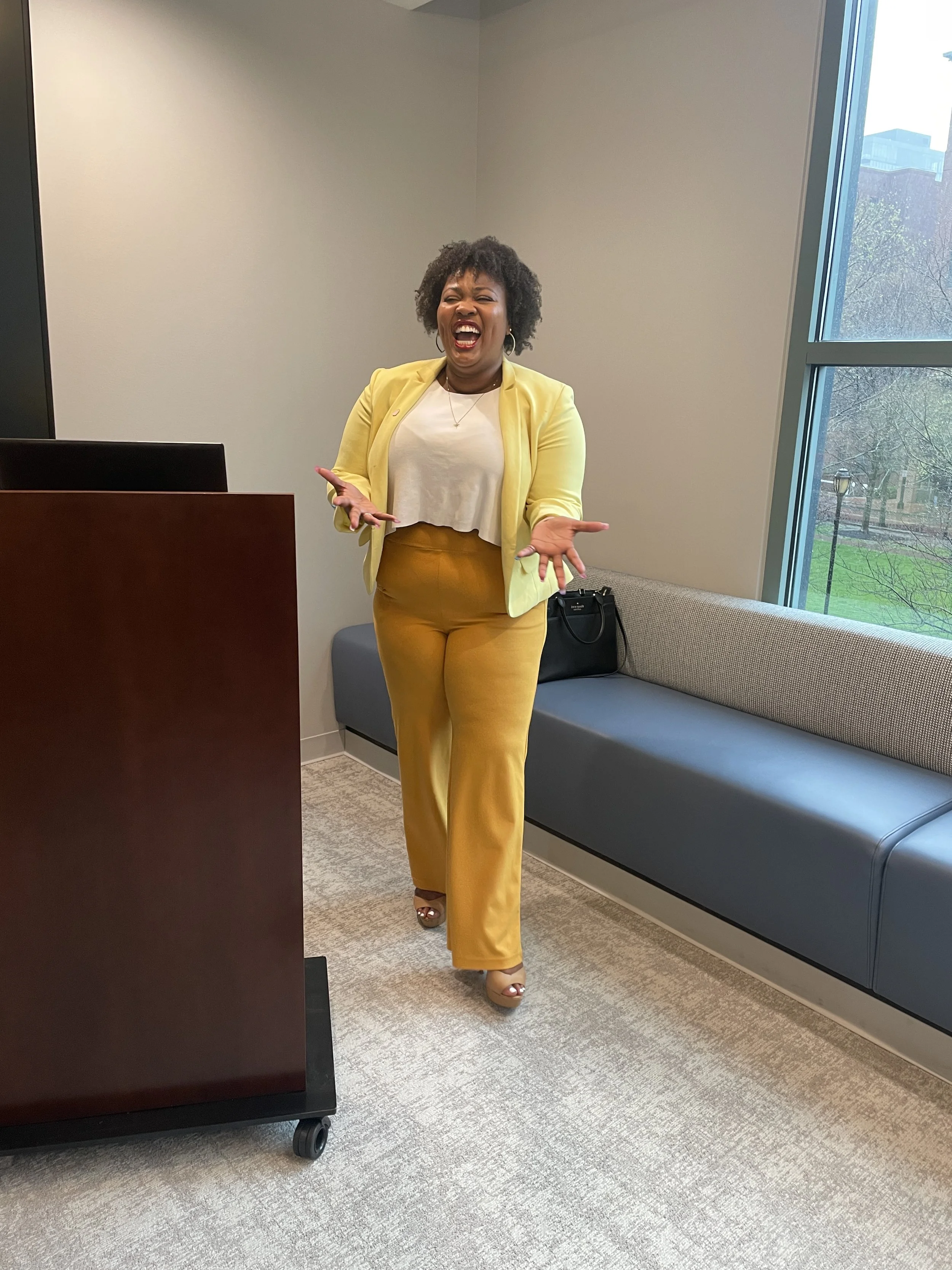 A woman in a yellow blazer and yellow pants laughing joyfully in a modern indoor setting with large windows and a grey bench.