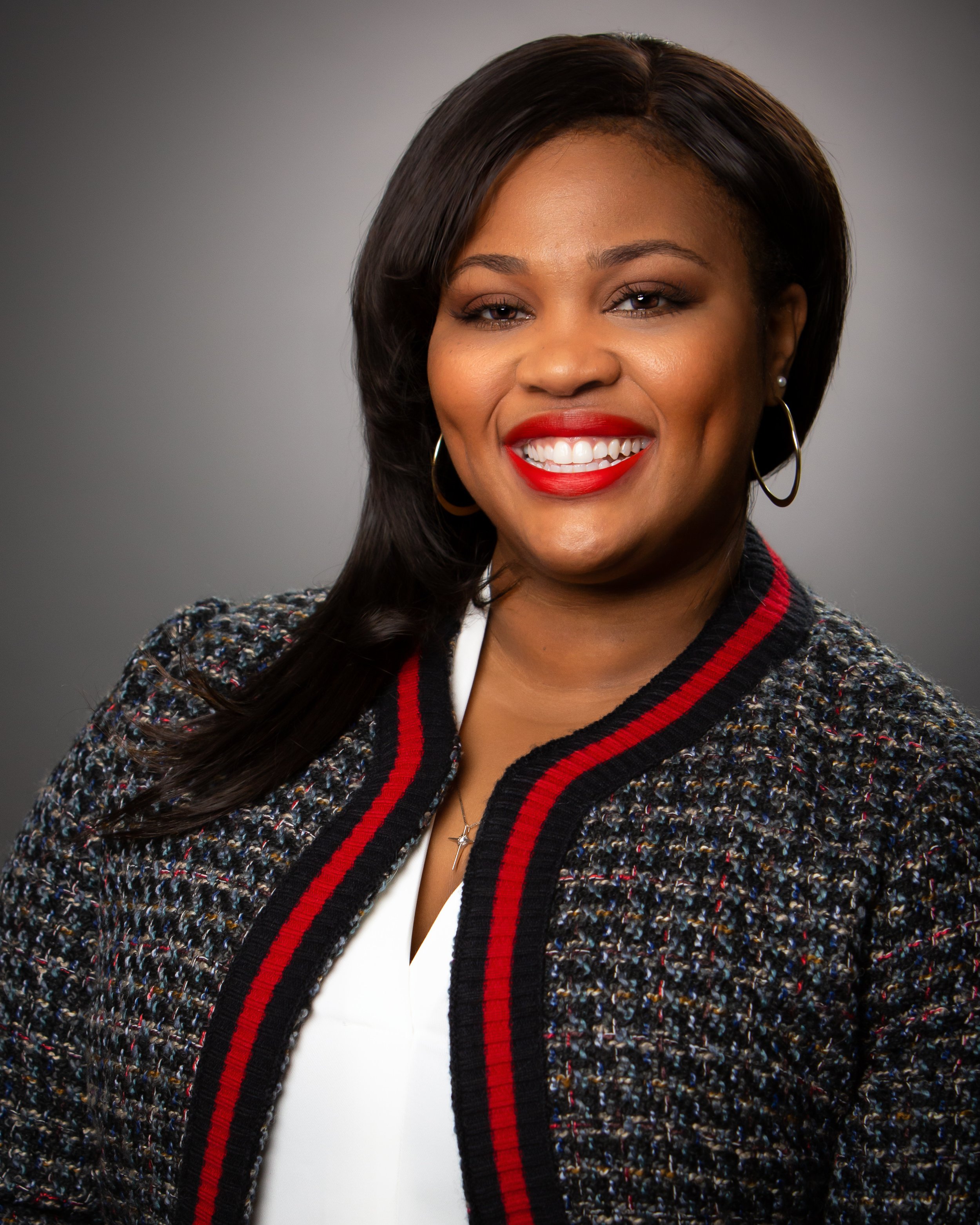 A smiling woman attorney with dark hair, wearing earrings, a necklace, and a multicolored textured suit jacket with red and black trim, set against a neutral background.
