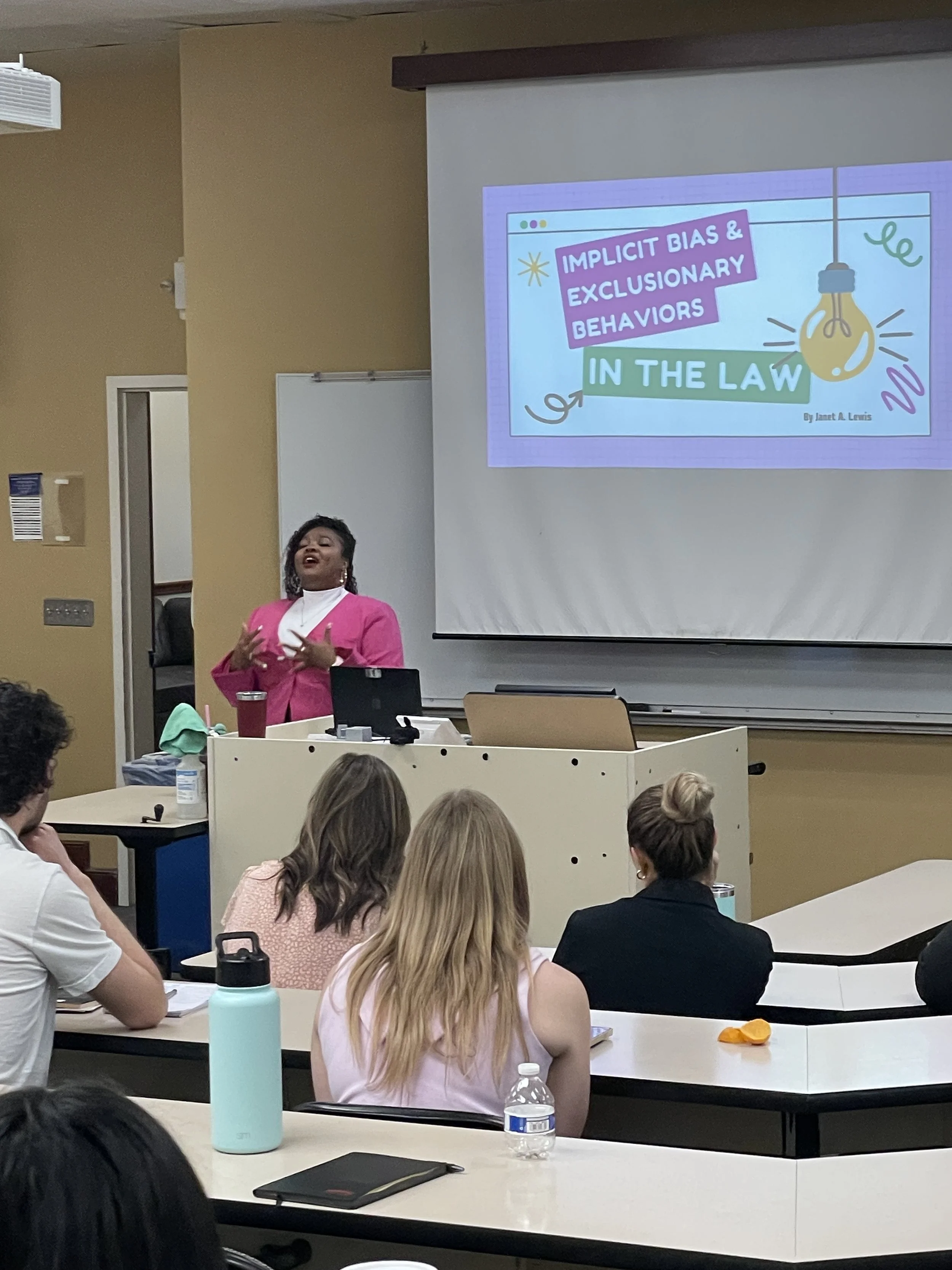 A woman wearing a pink blazer and white top giving a presentation in a classroom with a slide on the screen titled 'Implicit Bias & Exclusionary Behaviors in the Law', surrounded by colorful illustrations. Audience members are seated at tables, some taking notes or listening.