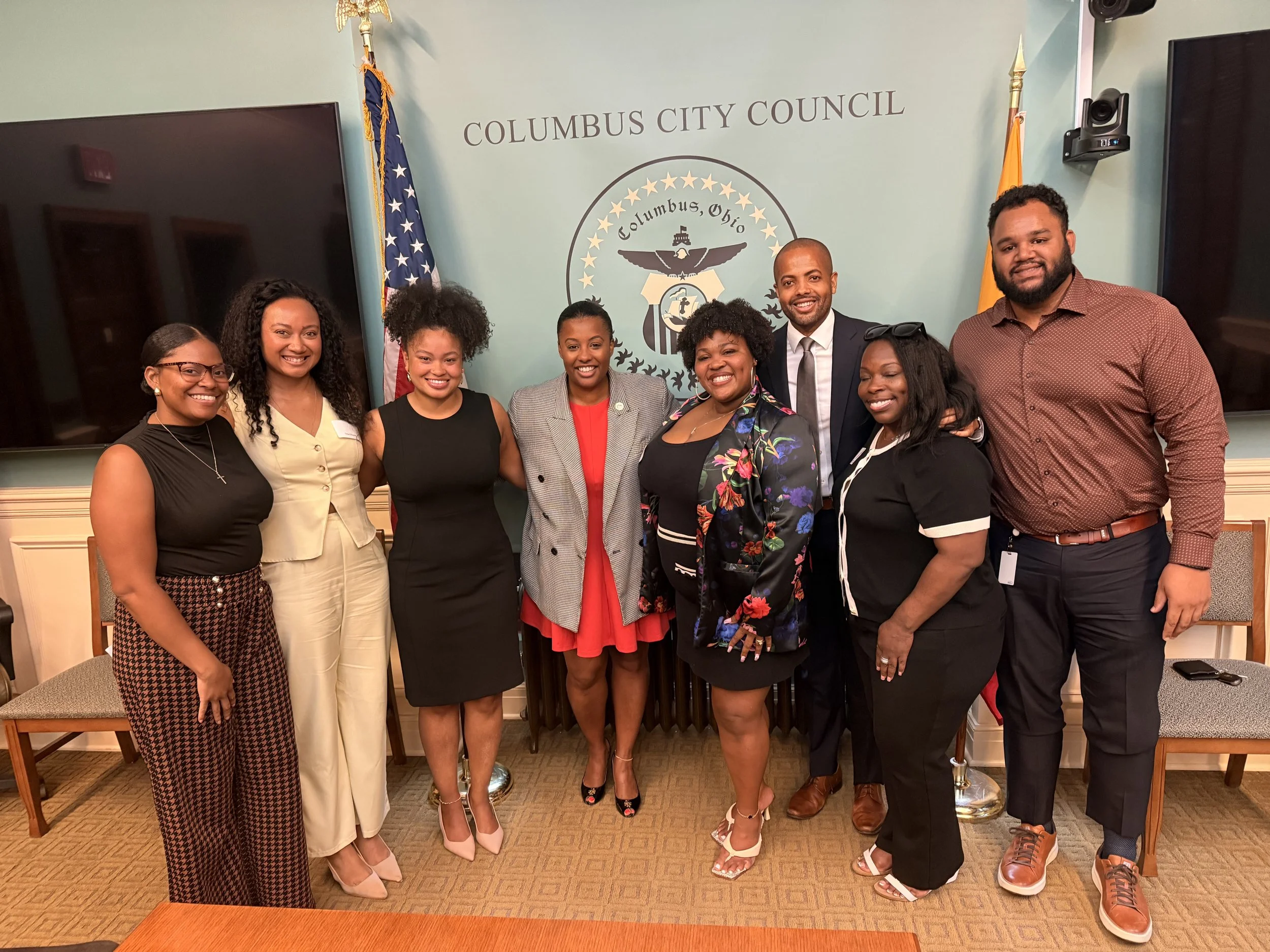 A group of nine diverse people, including women and men, standing together in front of a wall with 'Columbus City Council' written on it, posing for a photo. There are American flags and a seal on the wall.