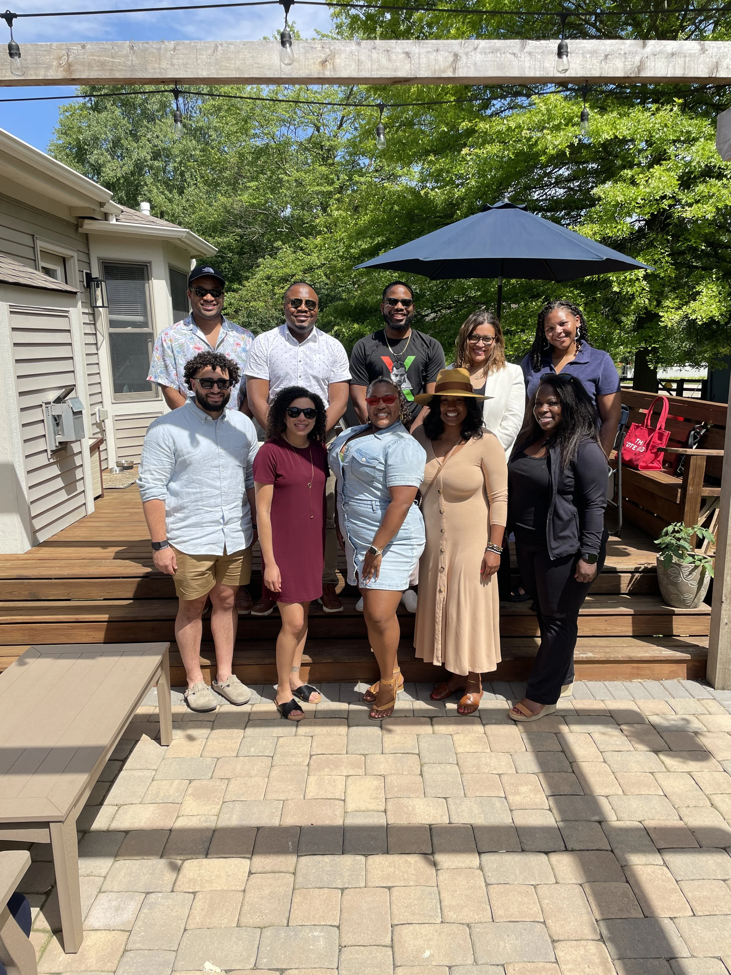 A group of eleven diverse people posing outdoors on a wooden deck under a string of outdoor lights, with lush green trees and a house in the background, sunny weather, and some wearing sunglasses.