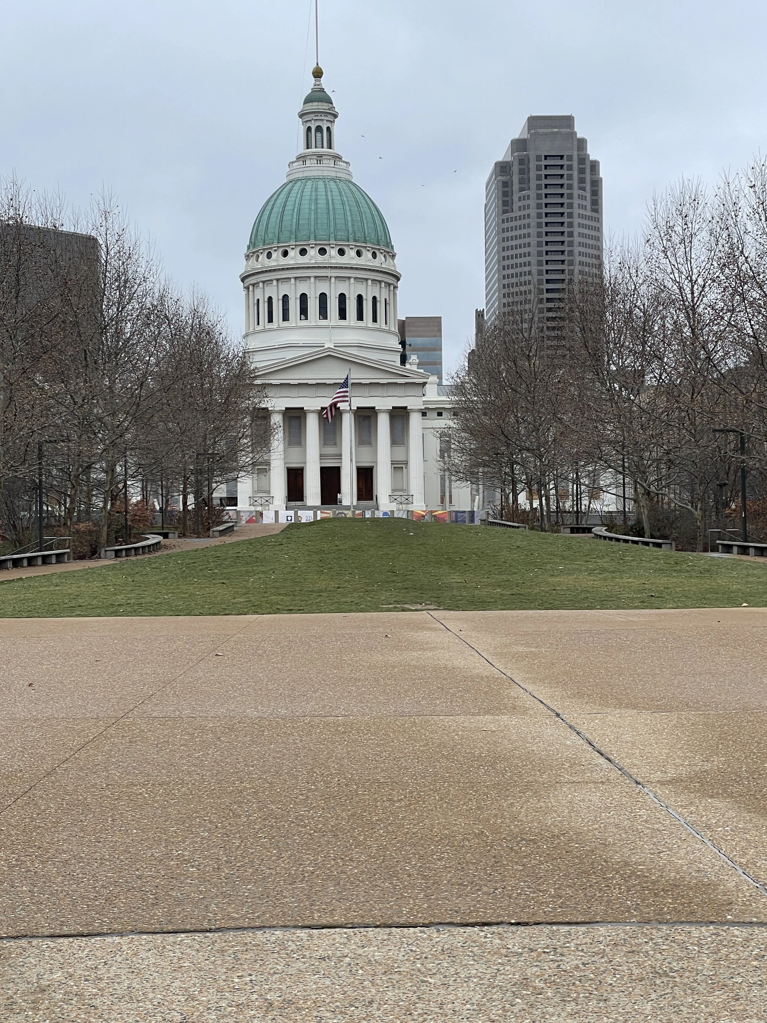 View of the Arkansas State Capitol with a green dome, white columns, and an American flag in front. Tall modern office buildings and leafless trees surround the capitol on a cloudy day.