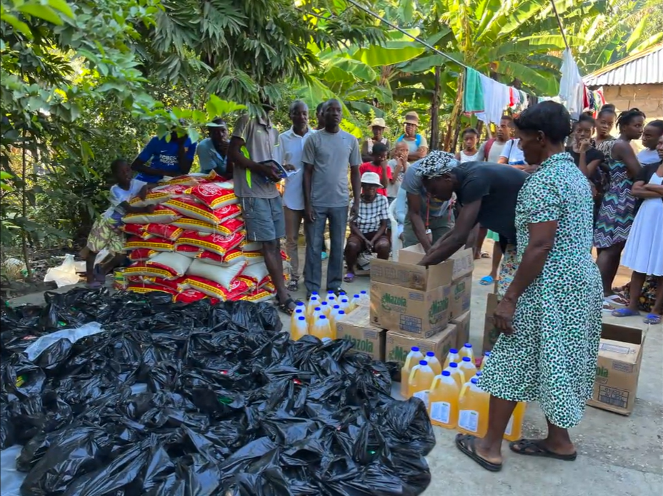 Community members distributing donated food supplies, bottled cooking oil, and packages in a tropical village.