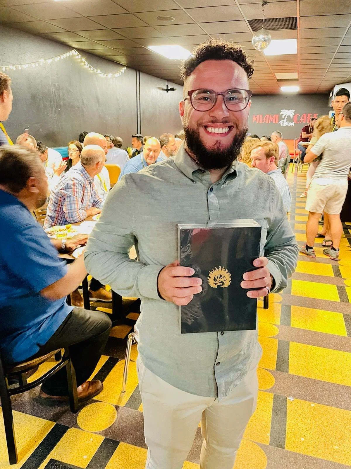 Smiling bearded man holding a black award plaque at a busy Miami Cafe restaurant