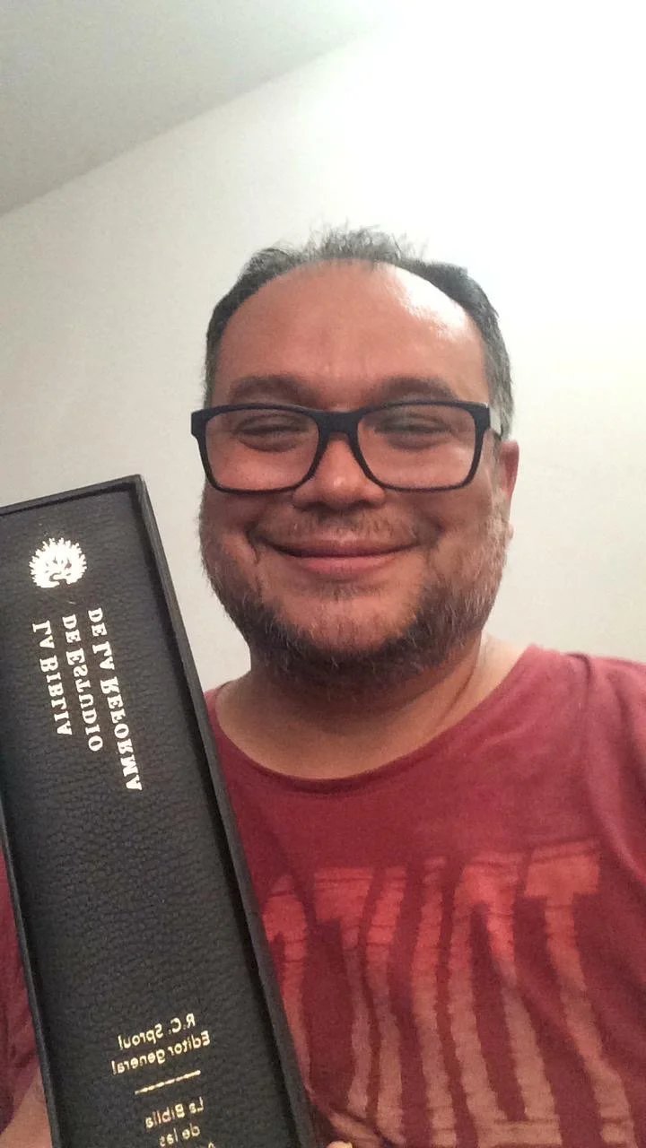 Smiling man wearing glasses and a red shirt holds a black award plaque with Spanish text.