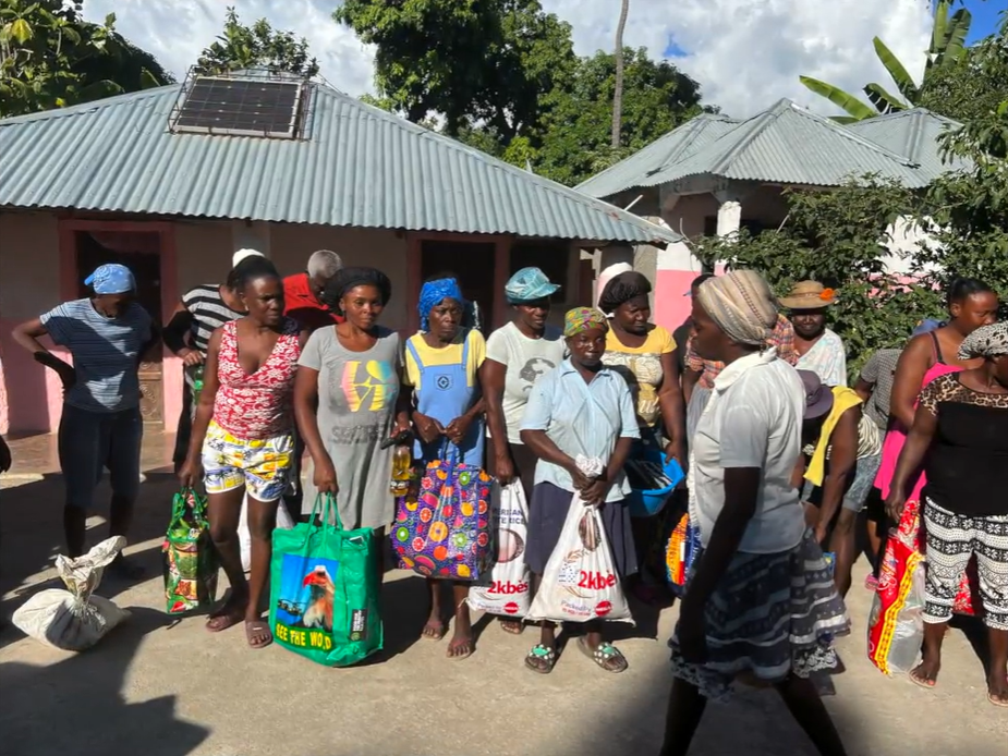 Group of people carrying colorful shopping bags gathered outside a corrugated metal-roofed building in a tropical village.