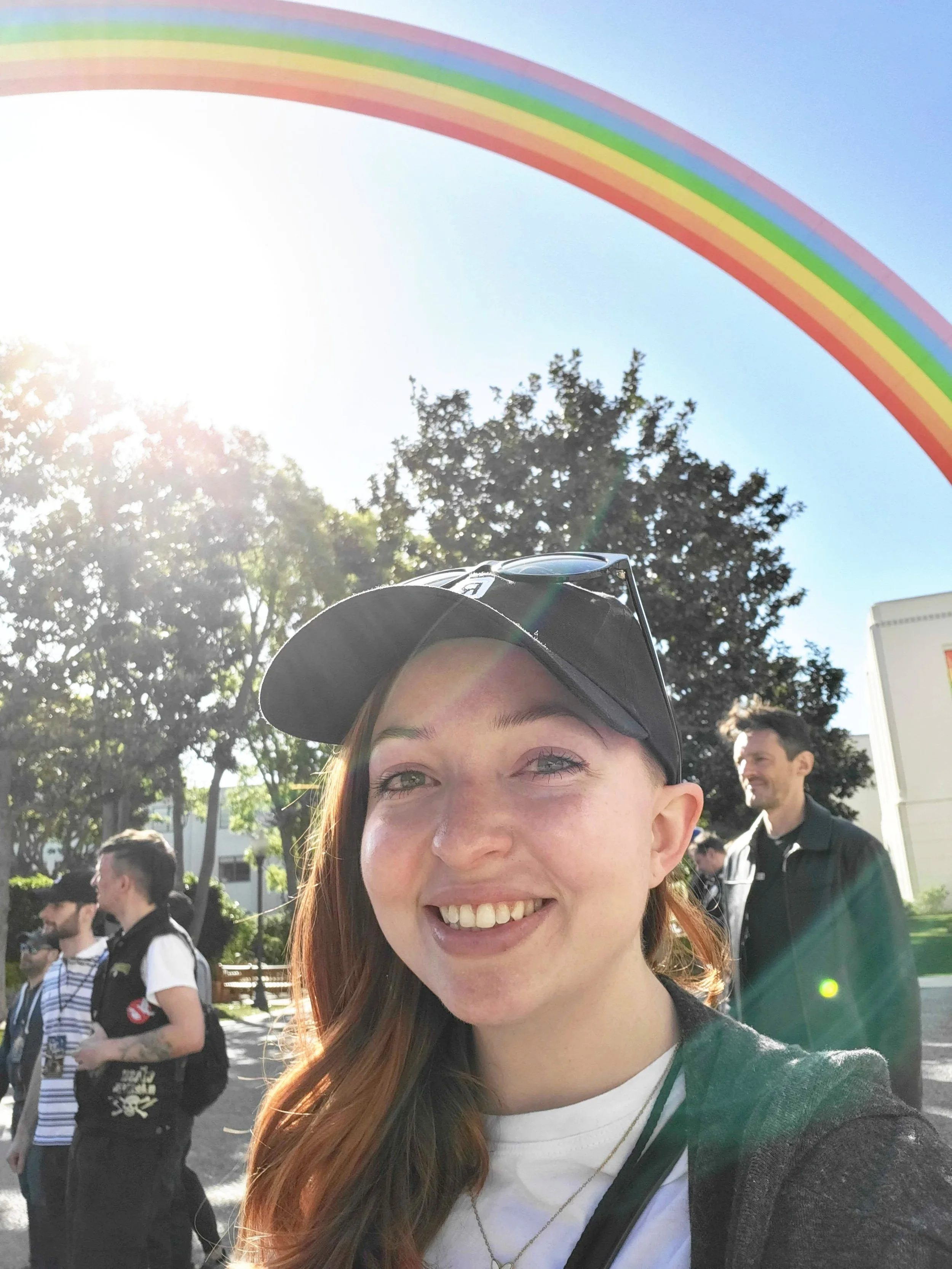 A young woman with red hair, smiling, wearing a black baseball cap and white t-shirt, standing outdoors in sunlight with trees and a rainbow decoration overhead, surrounded by other people.