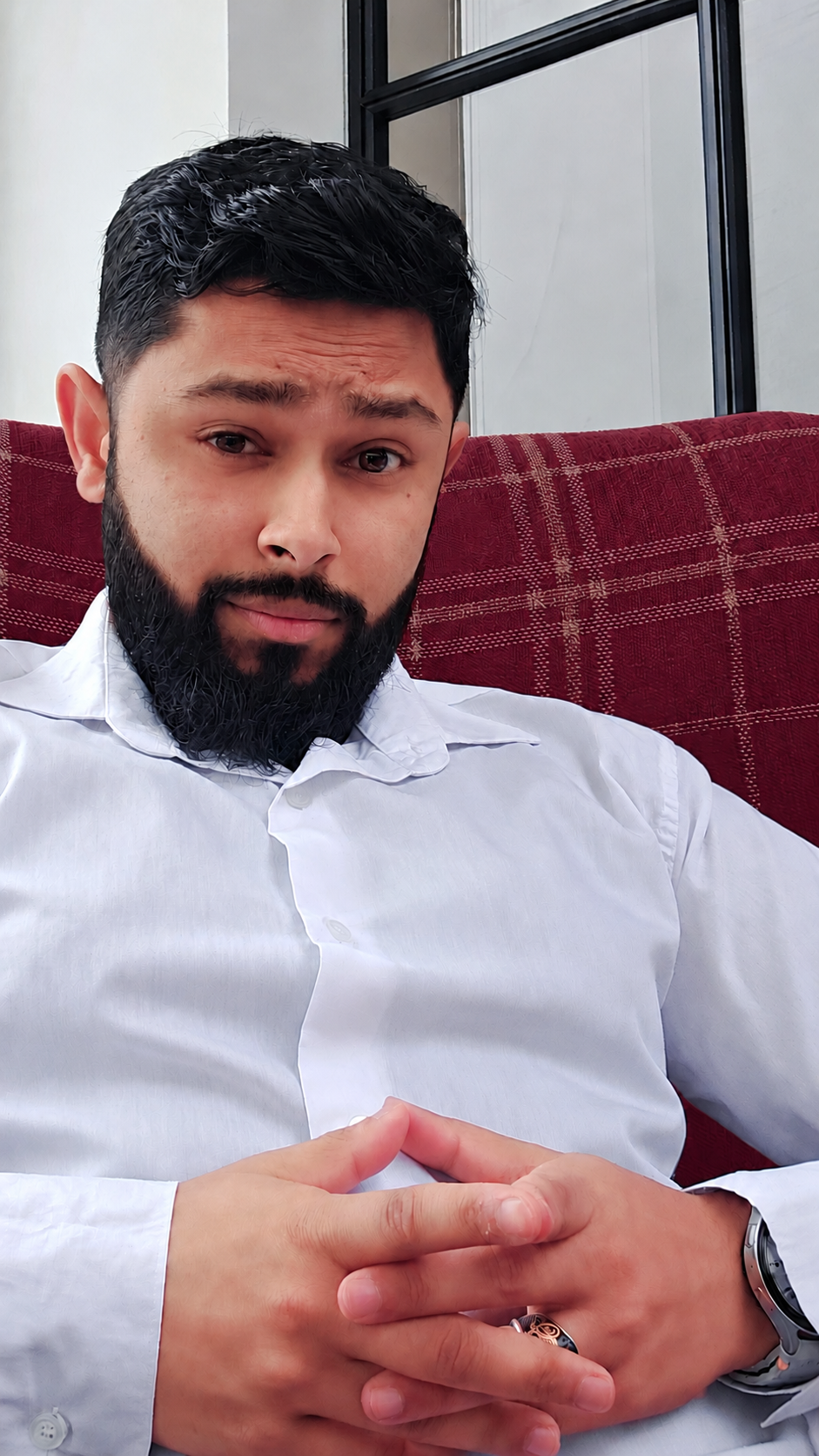 A man with a beard and dark hair, wearing a white button-up shirt, sitting on a red checkered sofa with his hands clasped together. He is in an indoor space near a window with a black frame.