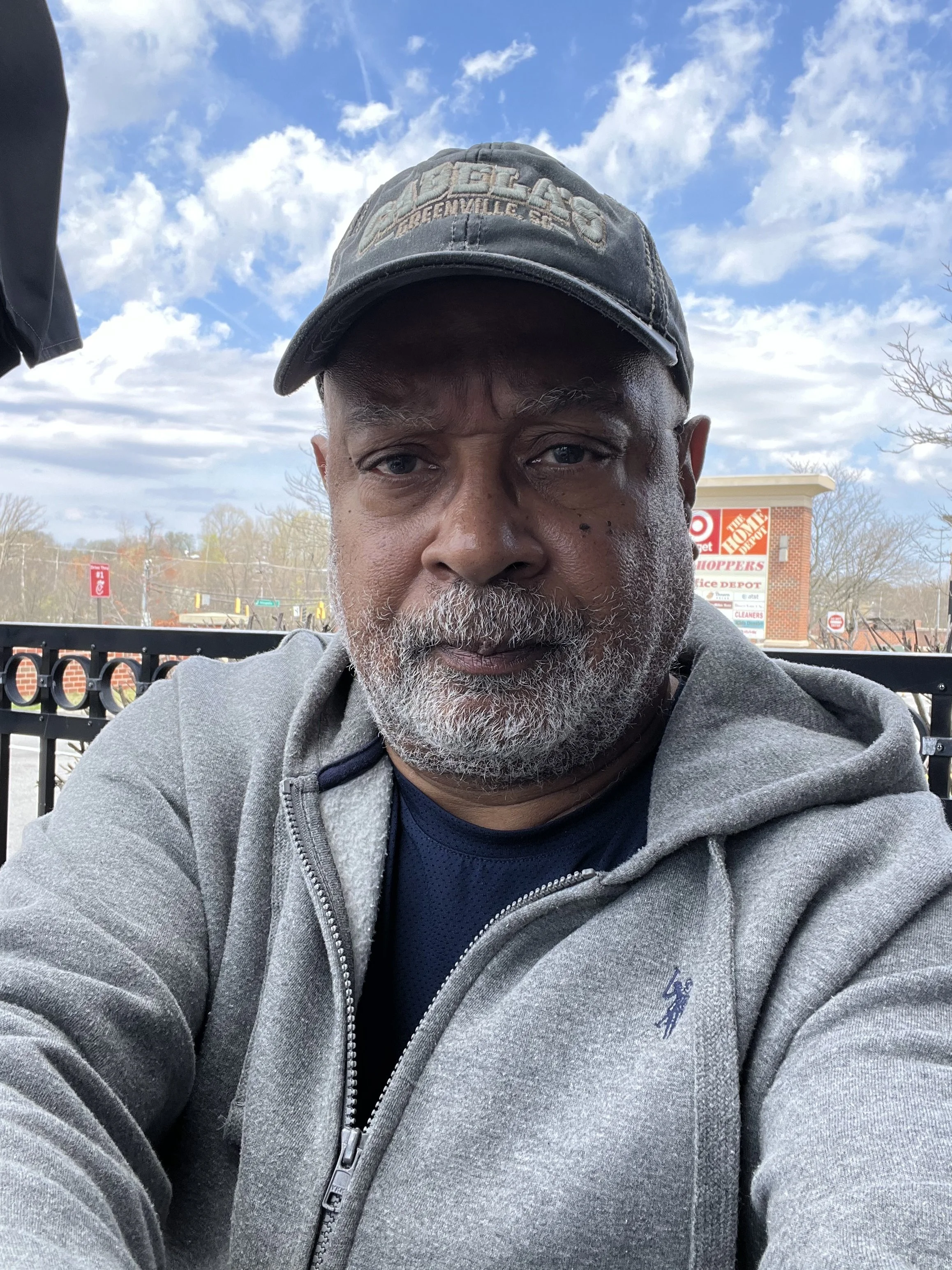 Close-up selfie of a bearded man wearing a black baseball cap and gray hoodie outdoors under a blue sky.
