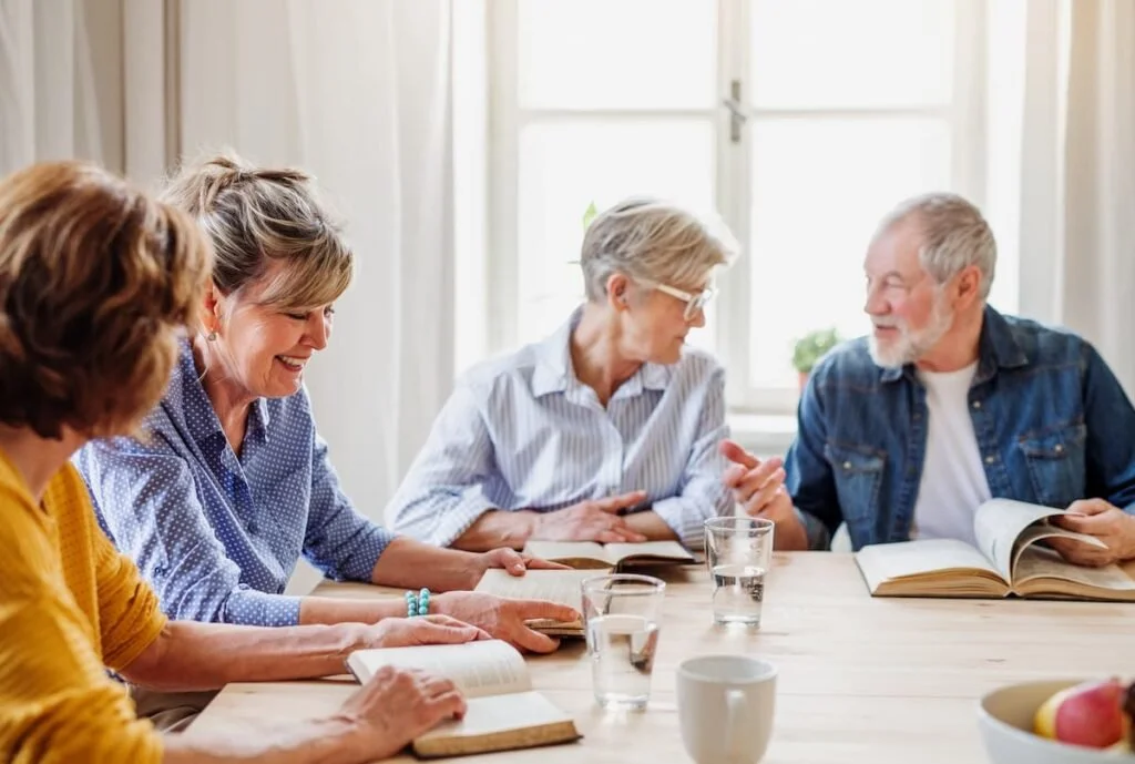 Four older adults discussing books around a table in a bright room.