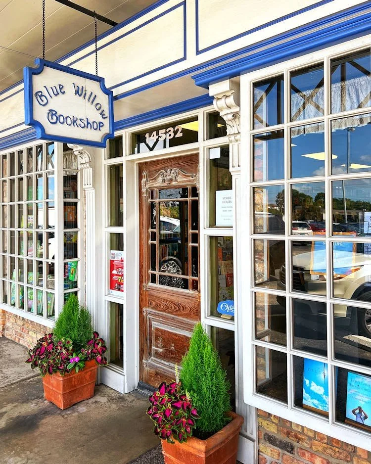 Blue Willow Bookshop storefront with ornate blue-and-white trim, wood door, book-filled windows, address 14532, and potted plants
