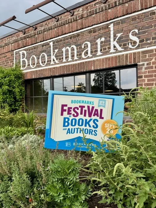Brick Bookmarks bookstore facade with a colorful Festival of Books and Authors sign surrounded by lush garden plants.
