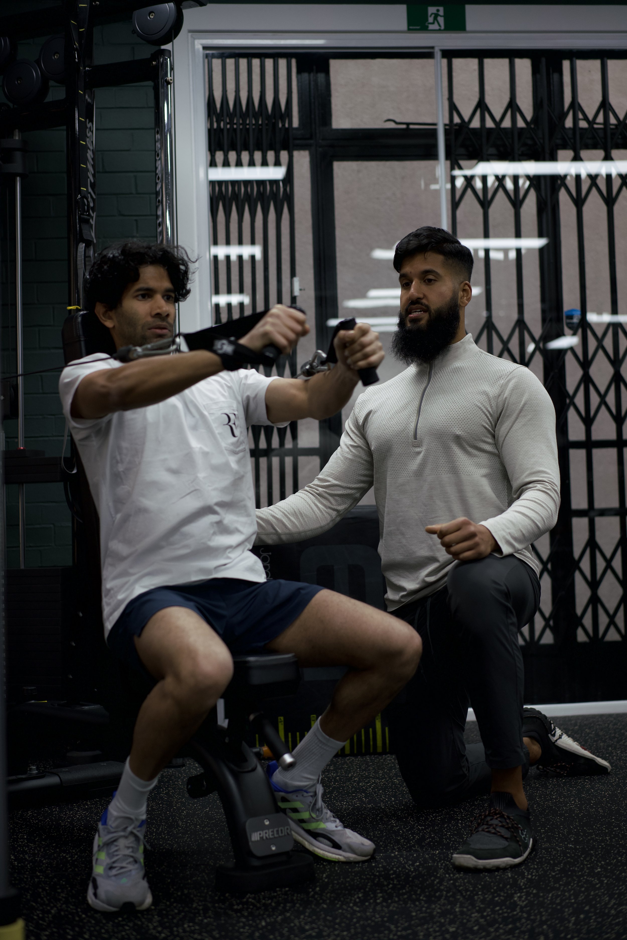 A man with curly black hair and a white t-shirt is sitting on a stationary exercise bike in a gym, while another man with a beard and gray long sleeve shirt is kneeling beside him, monitoring his activity.