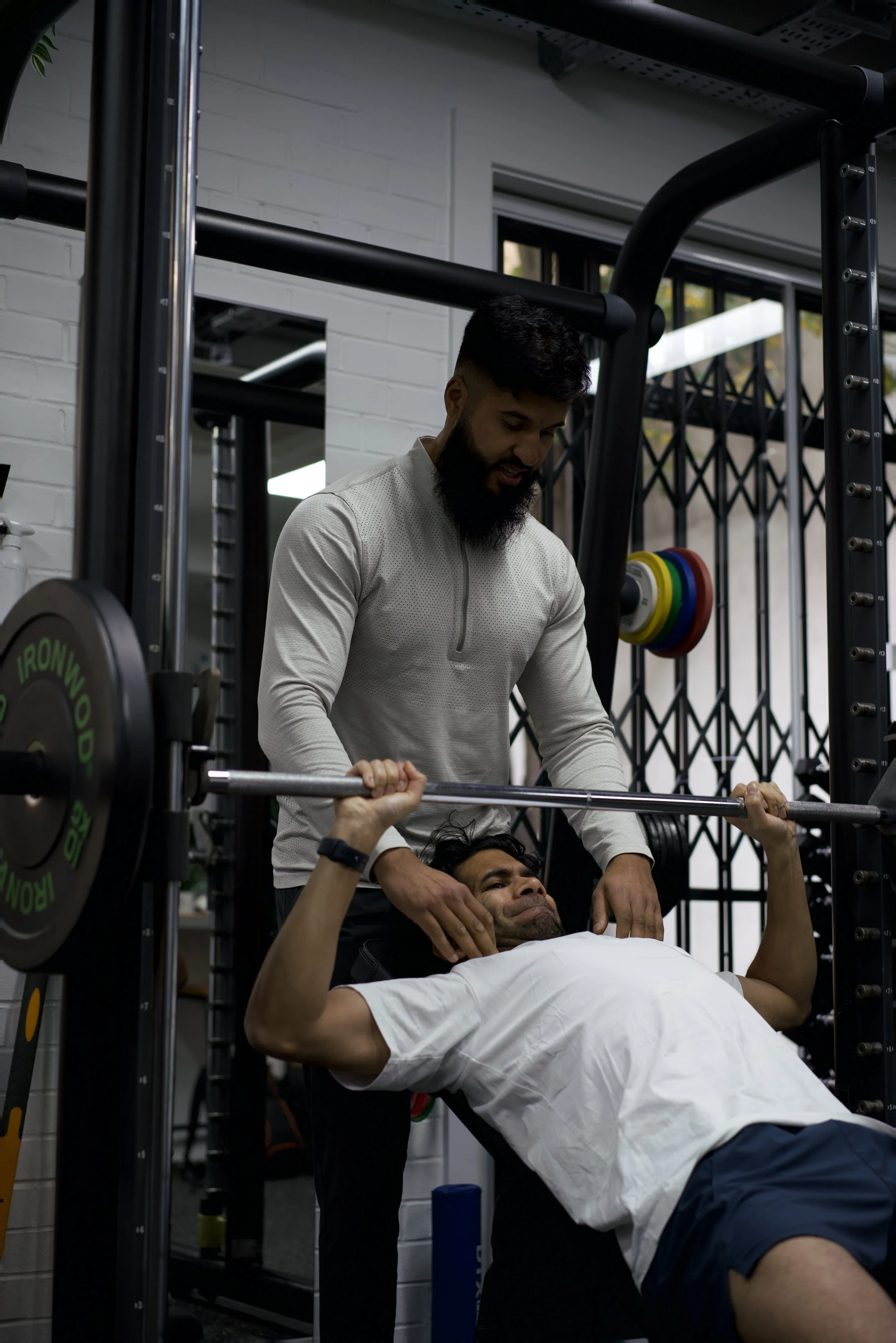 A man is lying on a bench press at the gym and lifting a barbell with assistance from a spotter. The spotter stands behind him, ready to assist if needed.