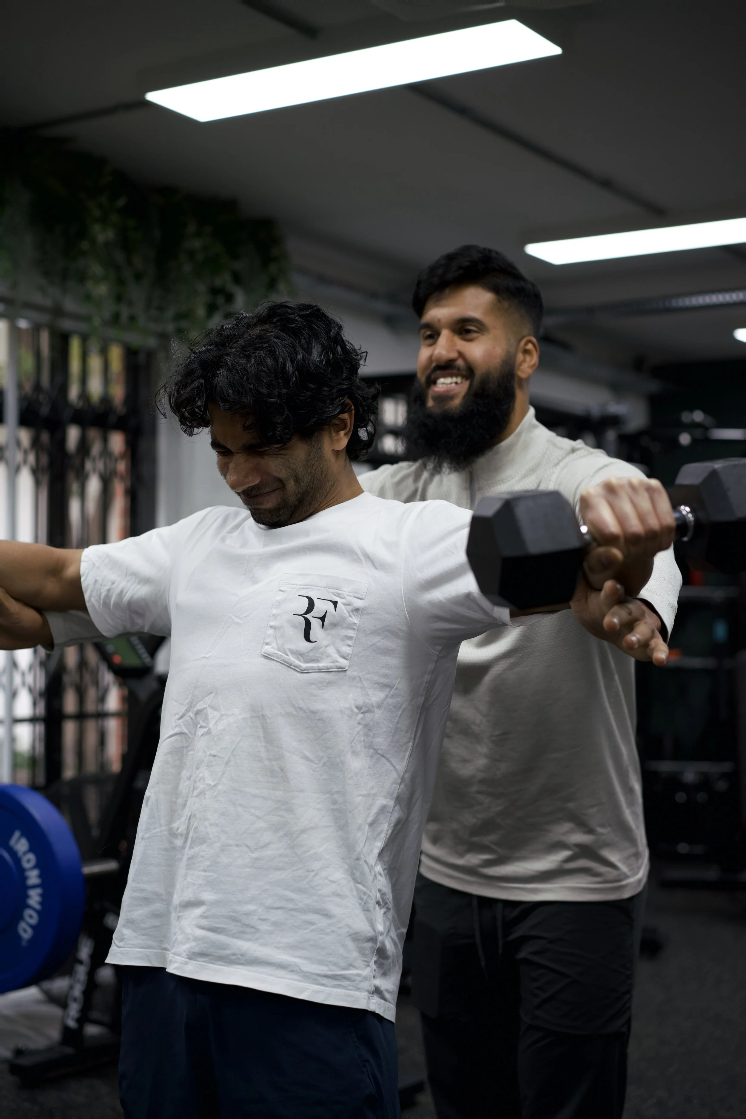 Two men working out in a gym, with one who is smiling and holding a dumbbell while the other assists him from behind.