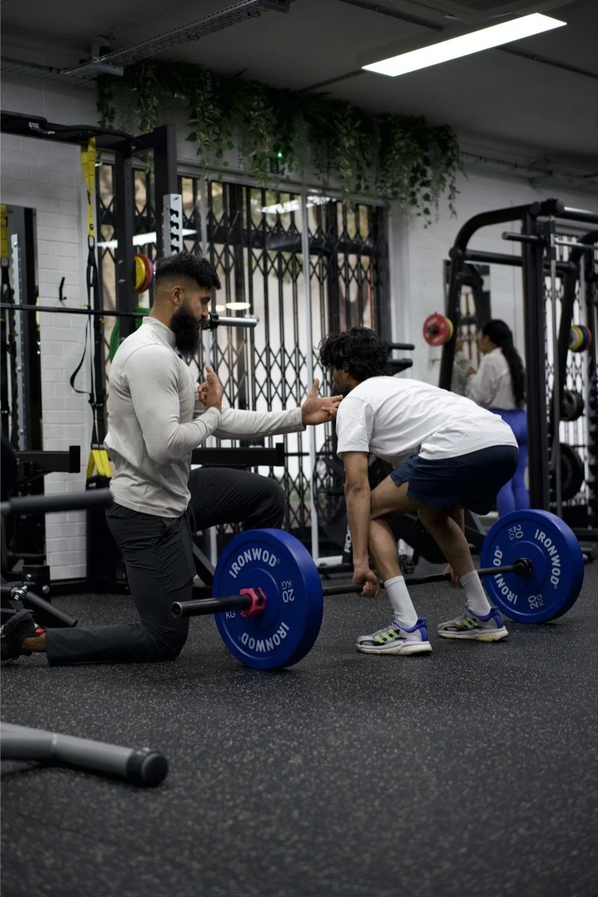 A personal trainer assists a young man with a deadlift exercise in a gym.