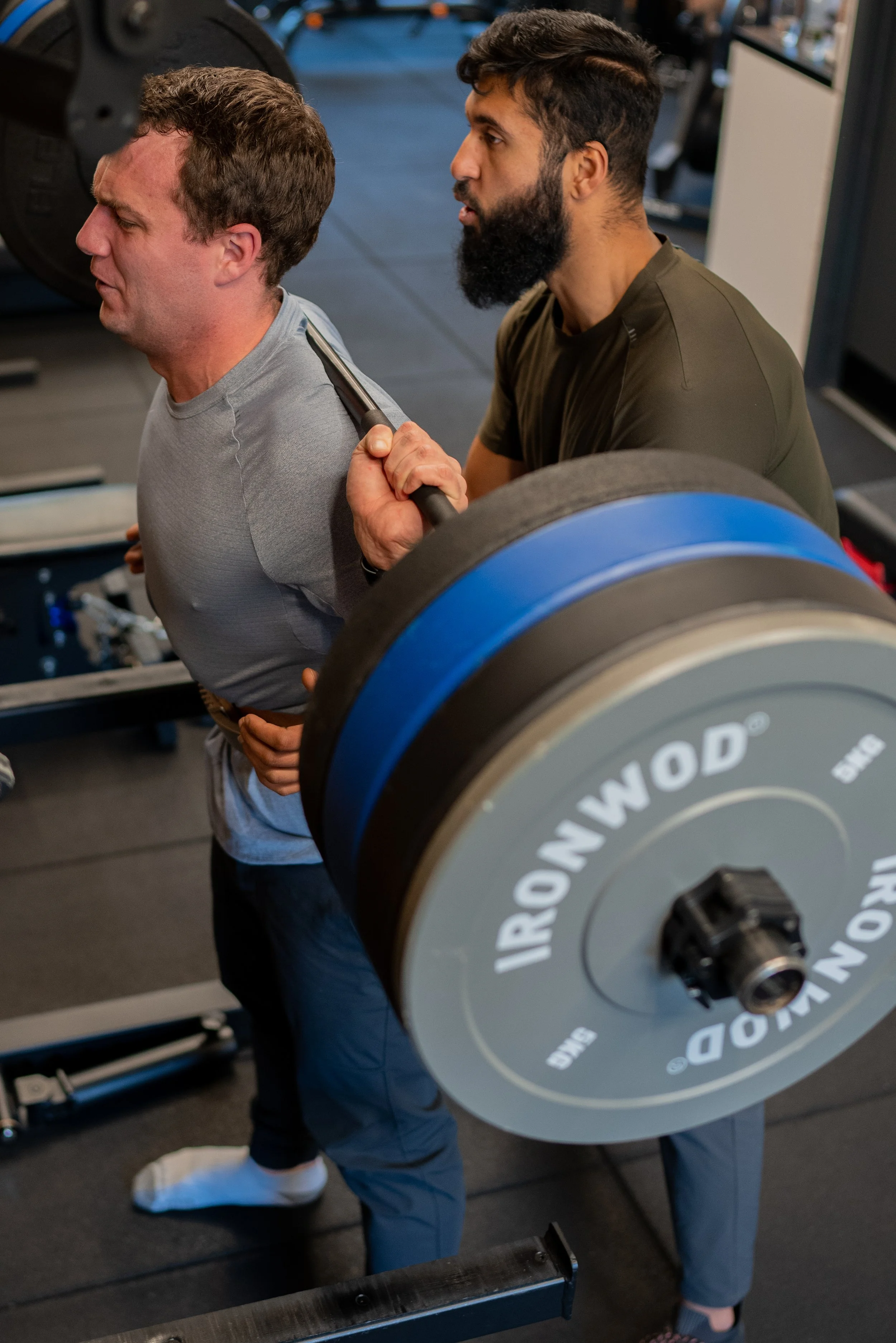A man lifting a barbell with weights at a gym, assisted by another man.