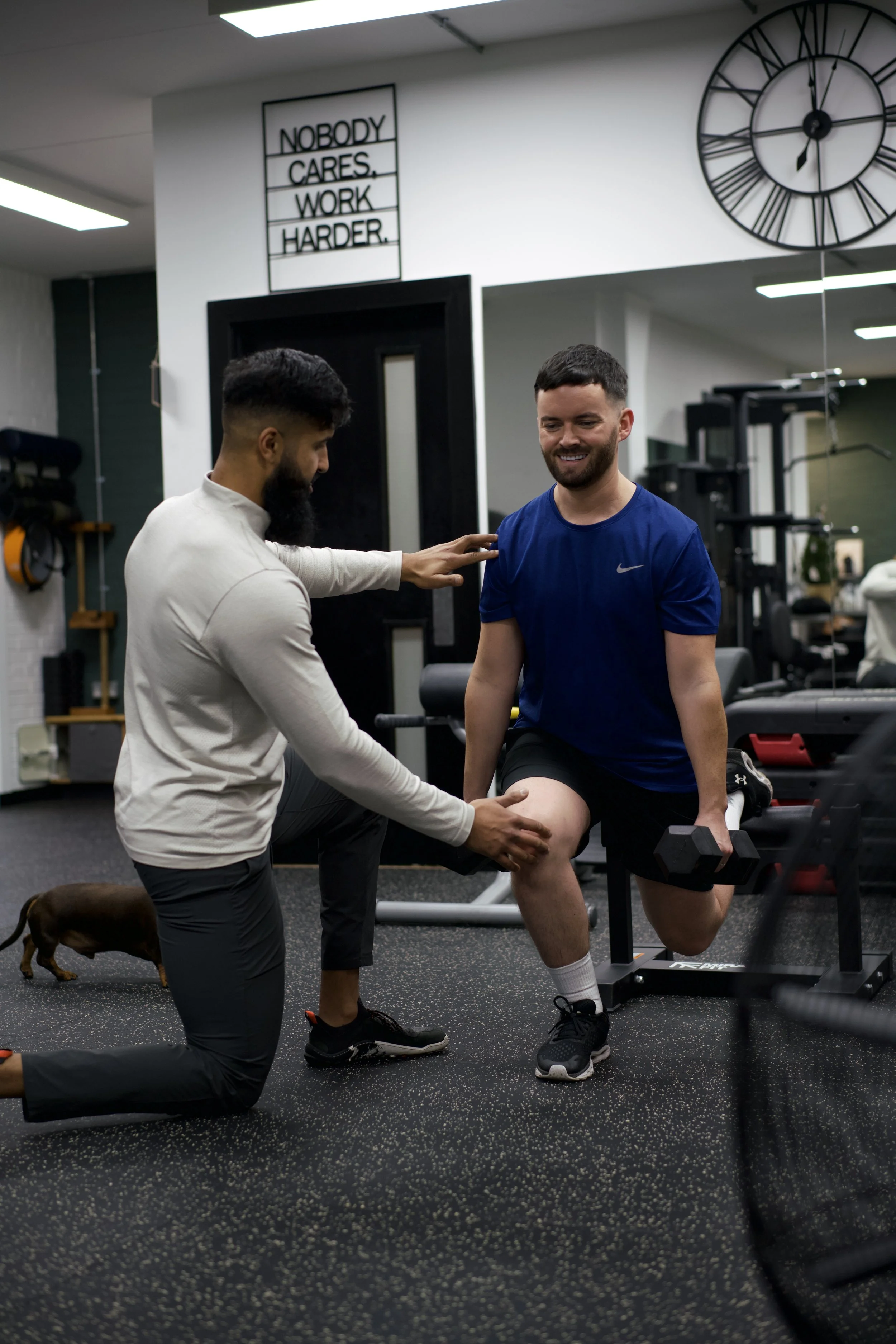 Two men helping an injured person during physical therapy in a gym, with one man kneeling and the other standing. A clock and motivational sign are on the wall behind them.