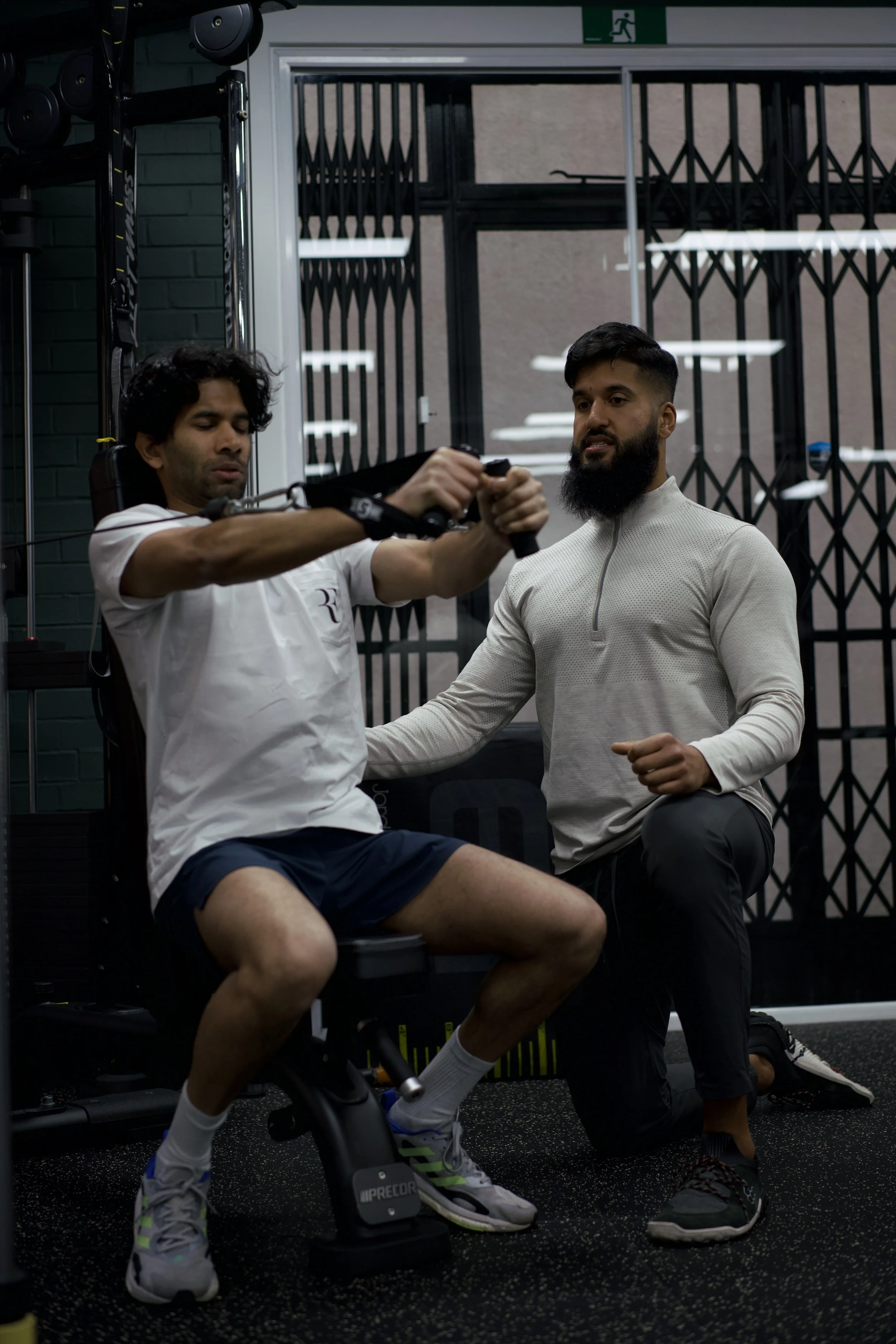 A man with a beard and a muscle shirt undergoing a physical therapy or training exercise with a trainer in a gym. The man is sitting on a chair, gripping a handle attached to a cable machine, while the trainer kneels beside him.