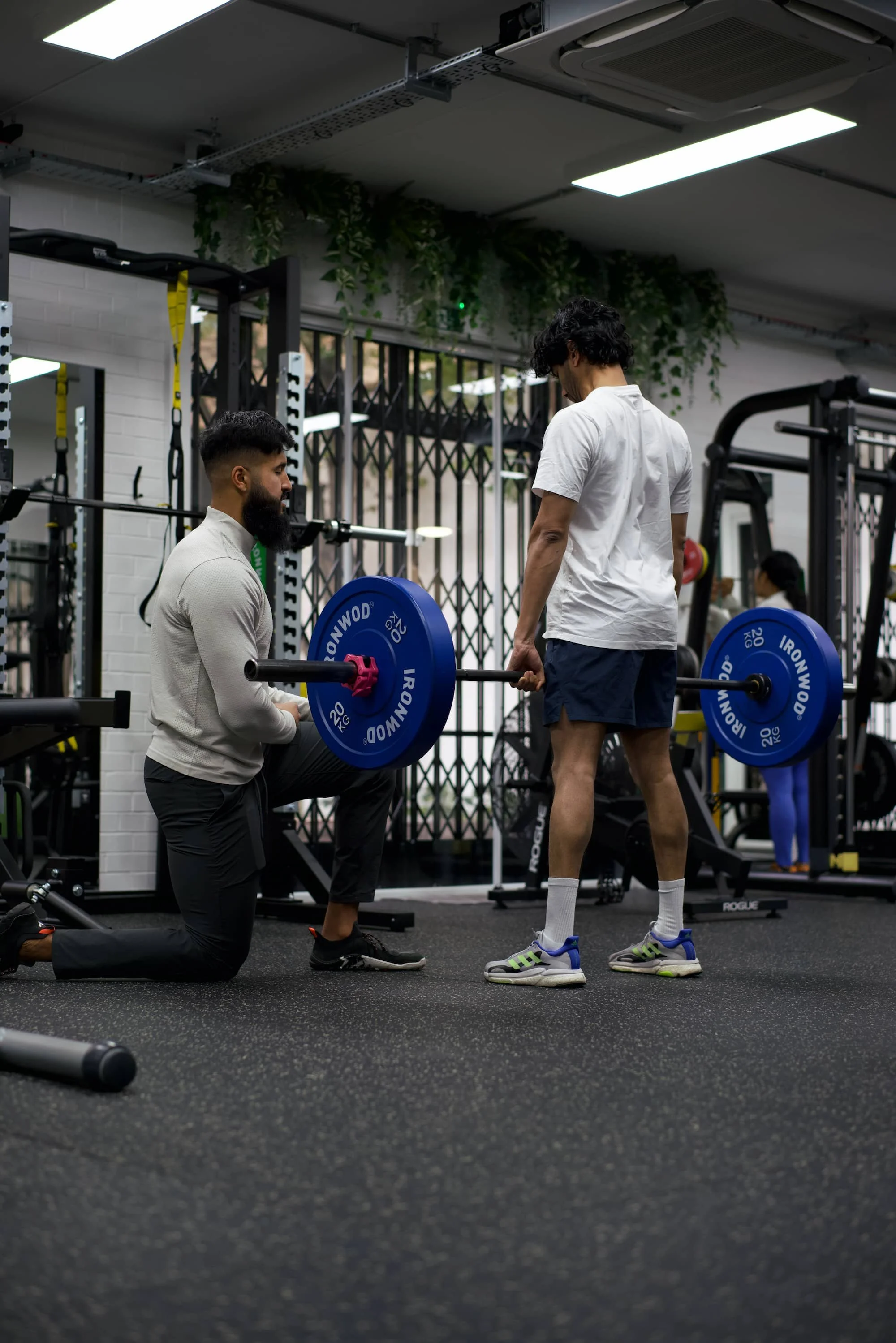 A trainer helping a man lift a barbell with blue weight plates in a gym, while another person watches. The gym has black flooring, fitness equipment, and large windows behind.
