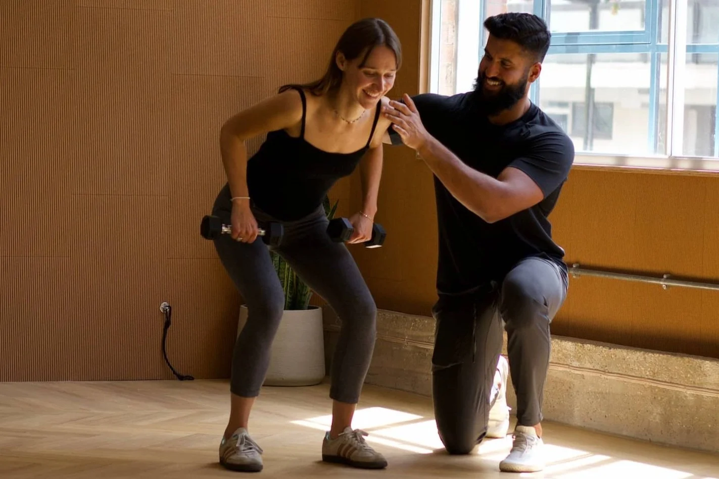 A woman exercises with dumbbells while a trainer offers encouragement indoors near a large window.