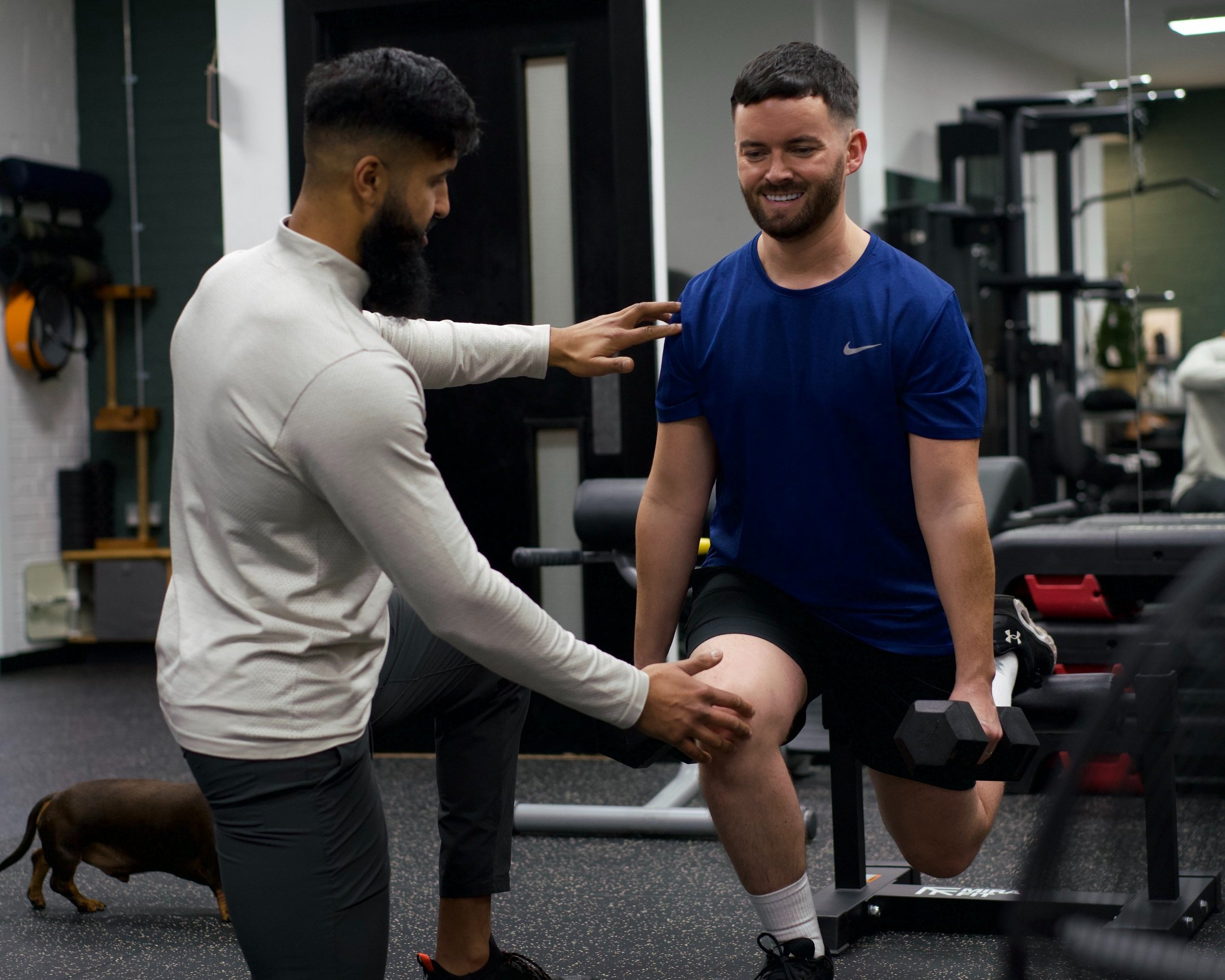 Two men are in a gym, one is kneeling with his arm extended on a bench, the other is standing, smiling, and holding a dumbbell in one hand and supporting the first man's knee with his other hand. A small brown dog is sitting on the floor nearby.