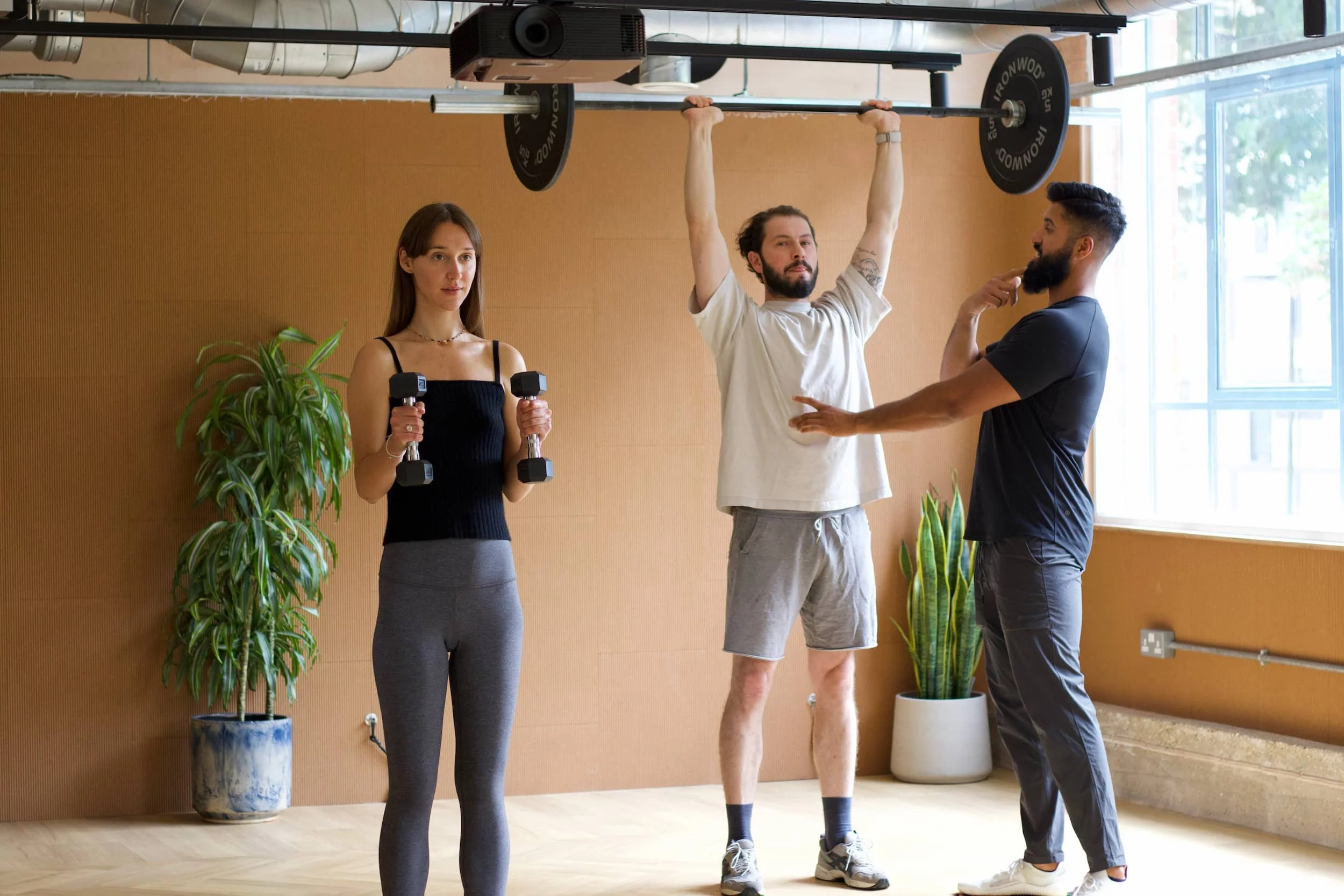 A woman in workout clothes holding dumbbells, a man lifting a barbell overhead with two women observing, in a gym with plants and large windows.