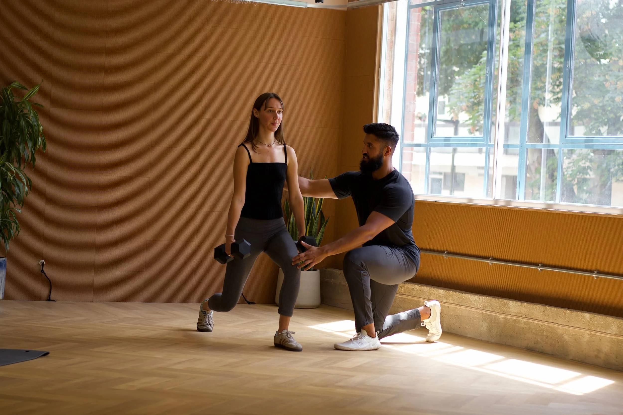 A man kneels on one knee providing support while a woman in workout clothes performs a lunge with a dumbbell in each hand in a bright room with large windows and wooden walls.