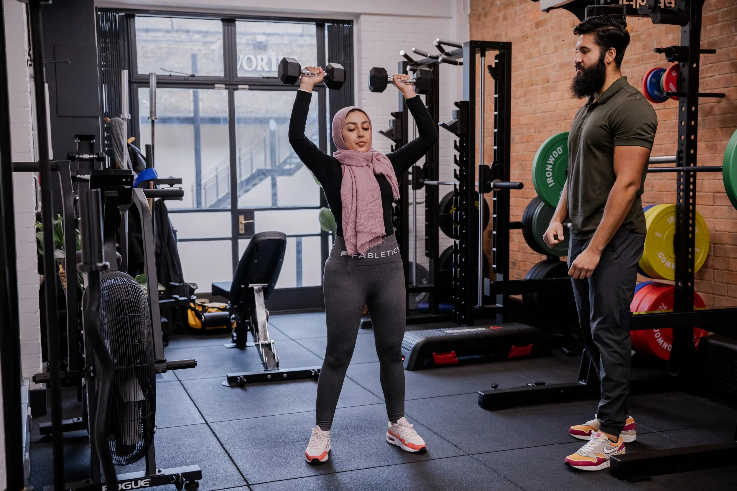 A woman lifting dumbbells during a workout in a gym, with a male trainer observing her.