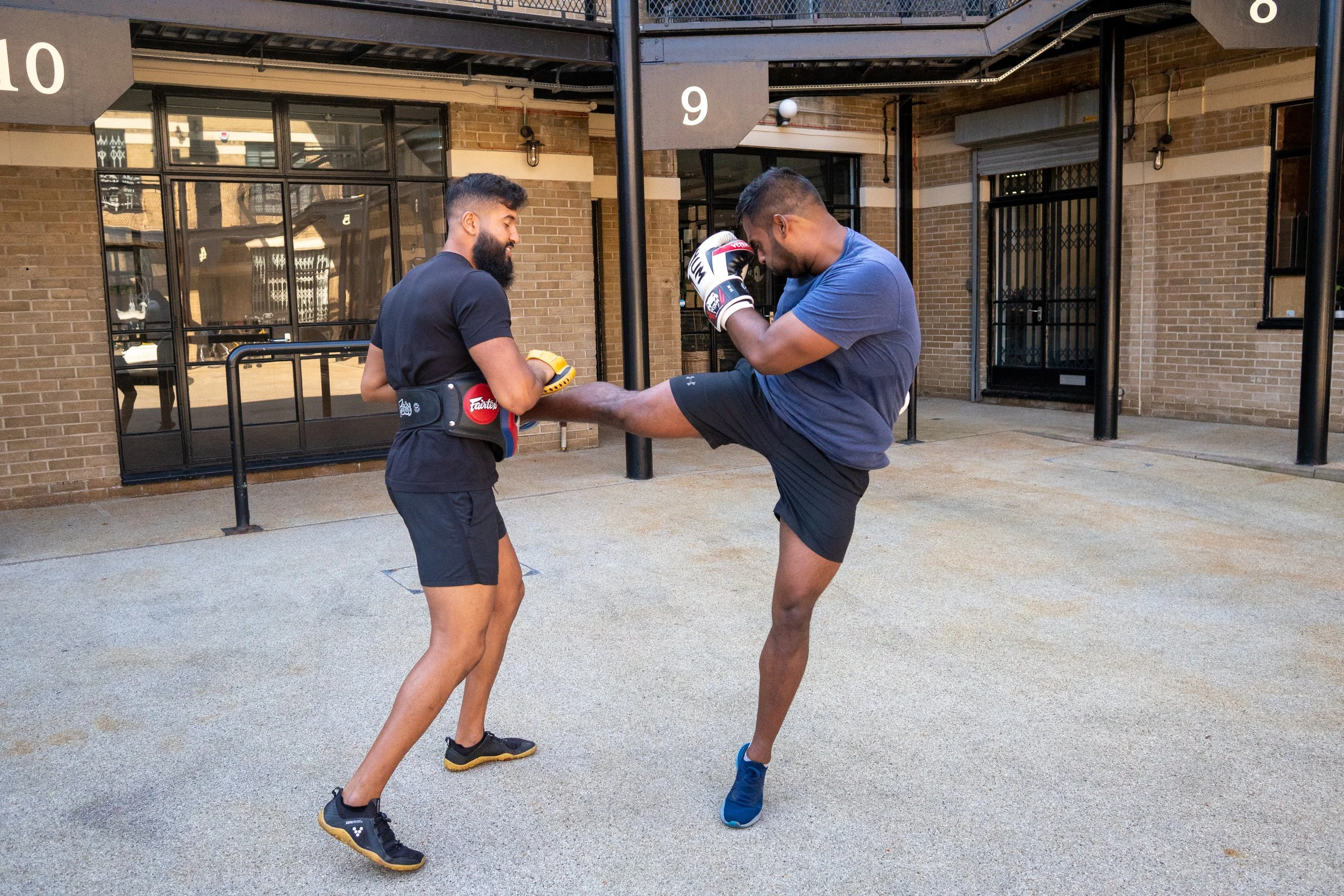 Two men practicing kickboxing outdoors in front of a brick building. One man has a beard and is holding yellow pads, while the other man, dressed in a blue t-shirt and shorts, is in a fighting stance and wearing boxing gloves.