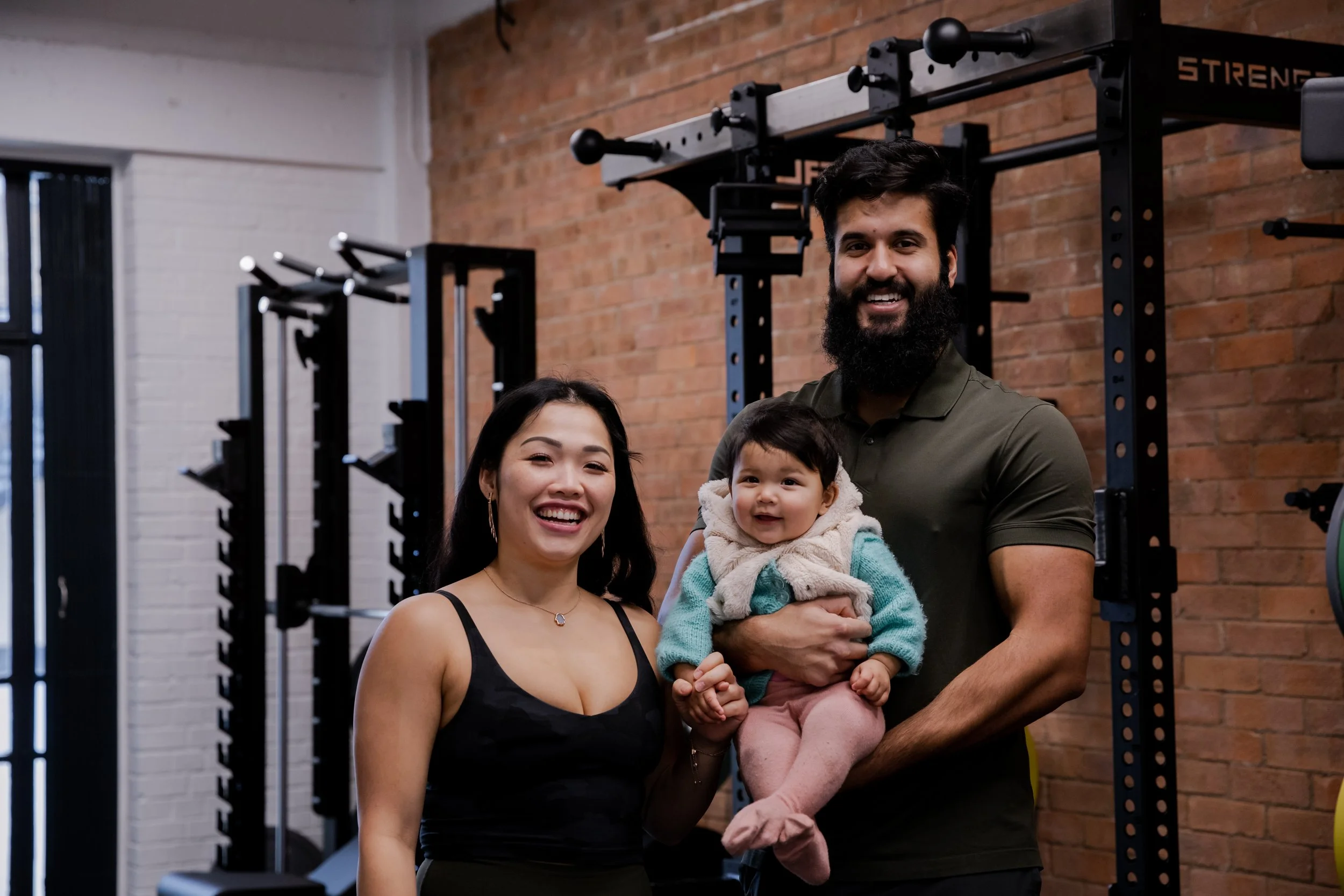 A smiling woman with long dark hair and a black top, a man with a beard and a green shirt holding a smiling baby girl in a gym with brick walls and workout equipment in the background.