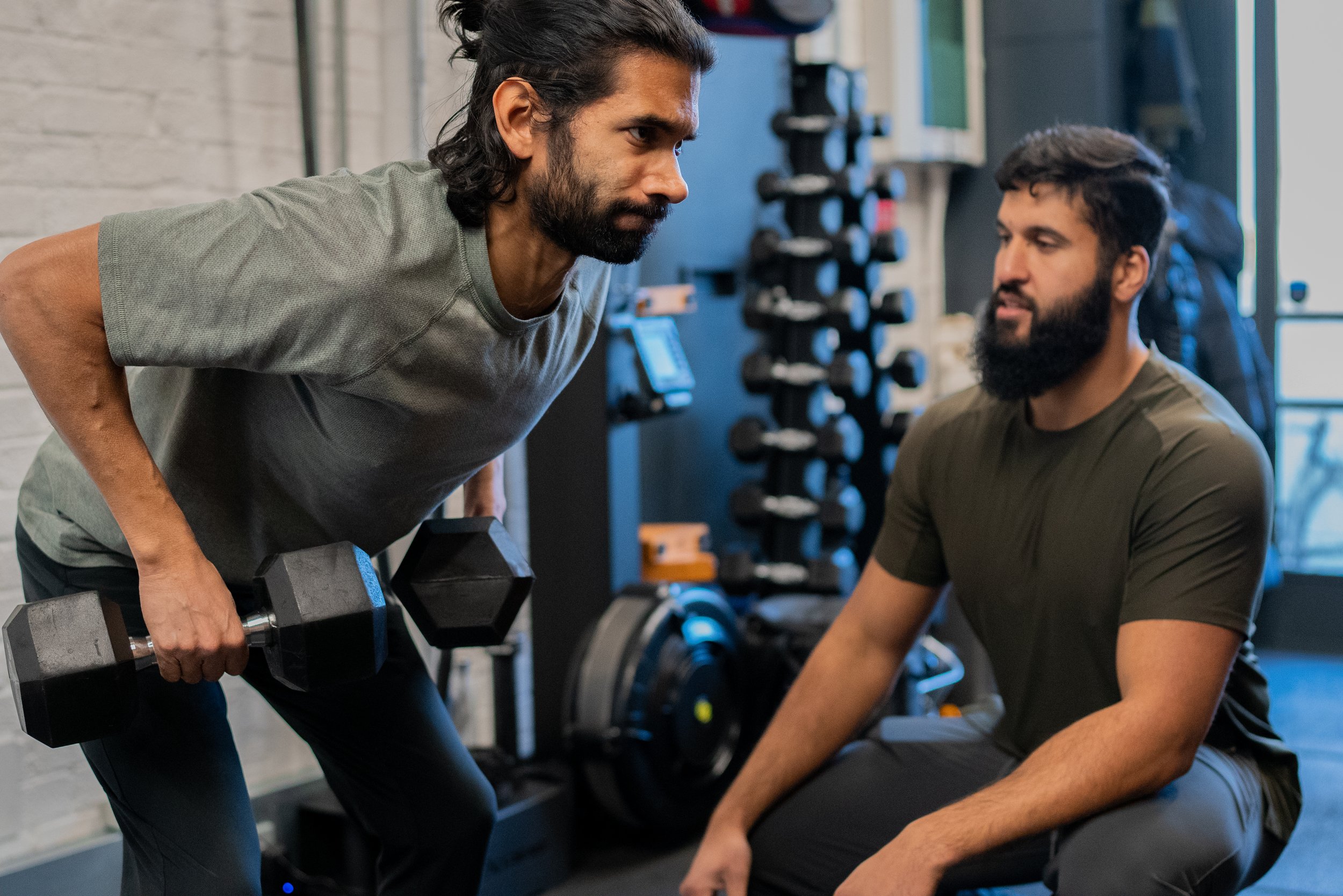 Two men working out in a gym. One man is lifting a dumbbell and the other man is sitting, possibly receiving guidance or encouragement.