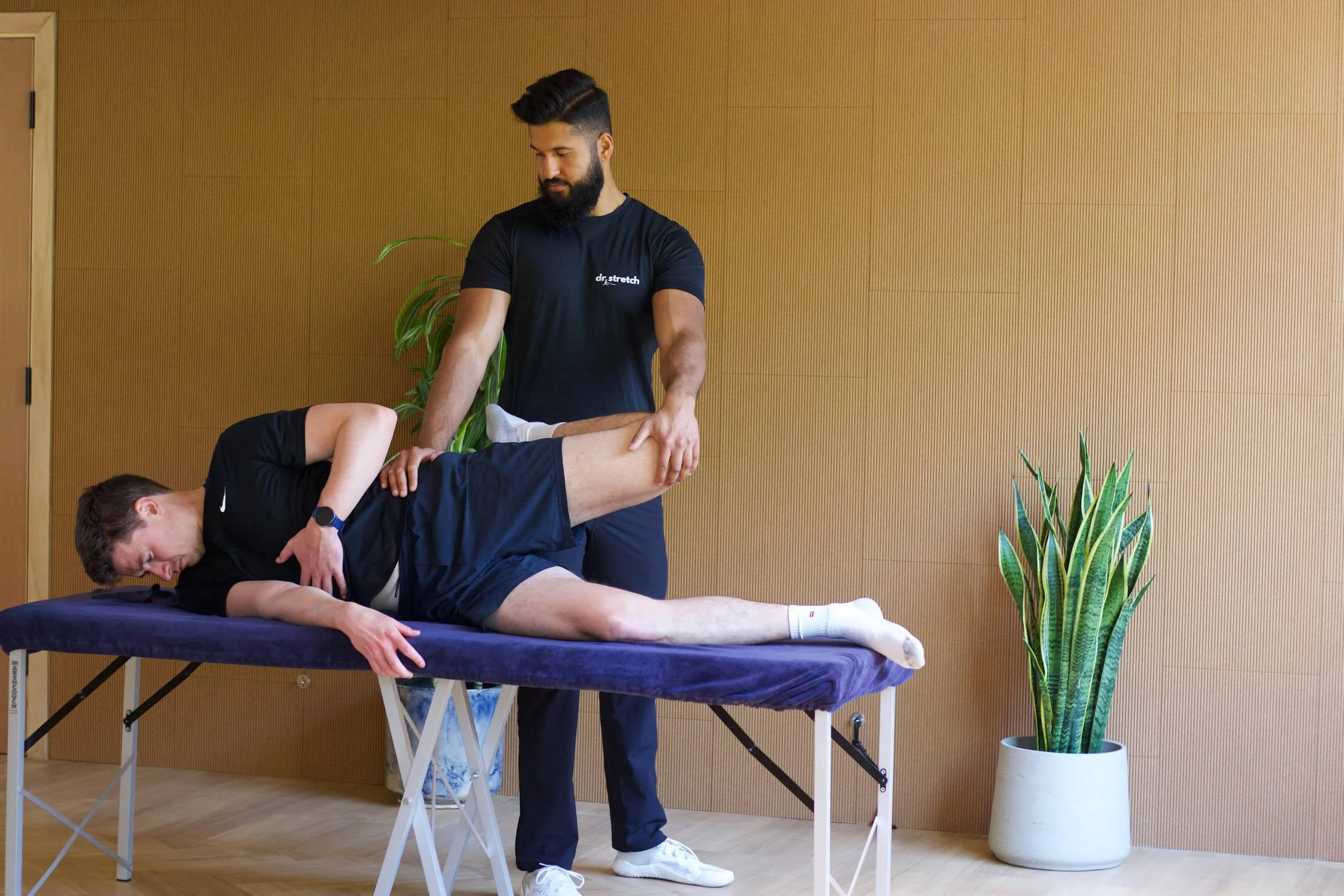 A man receives a physical therapy treatment from a professional who is stretching his leg on a massage table in a room with wooden paneled wall and a green plant in a white pot.