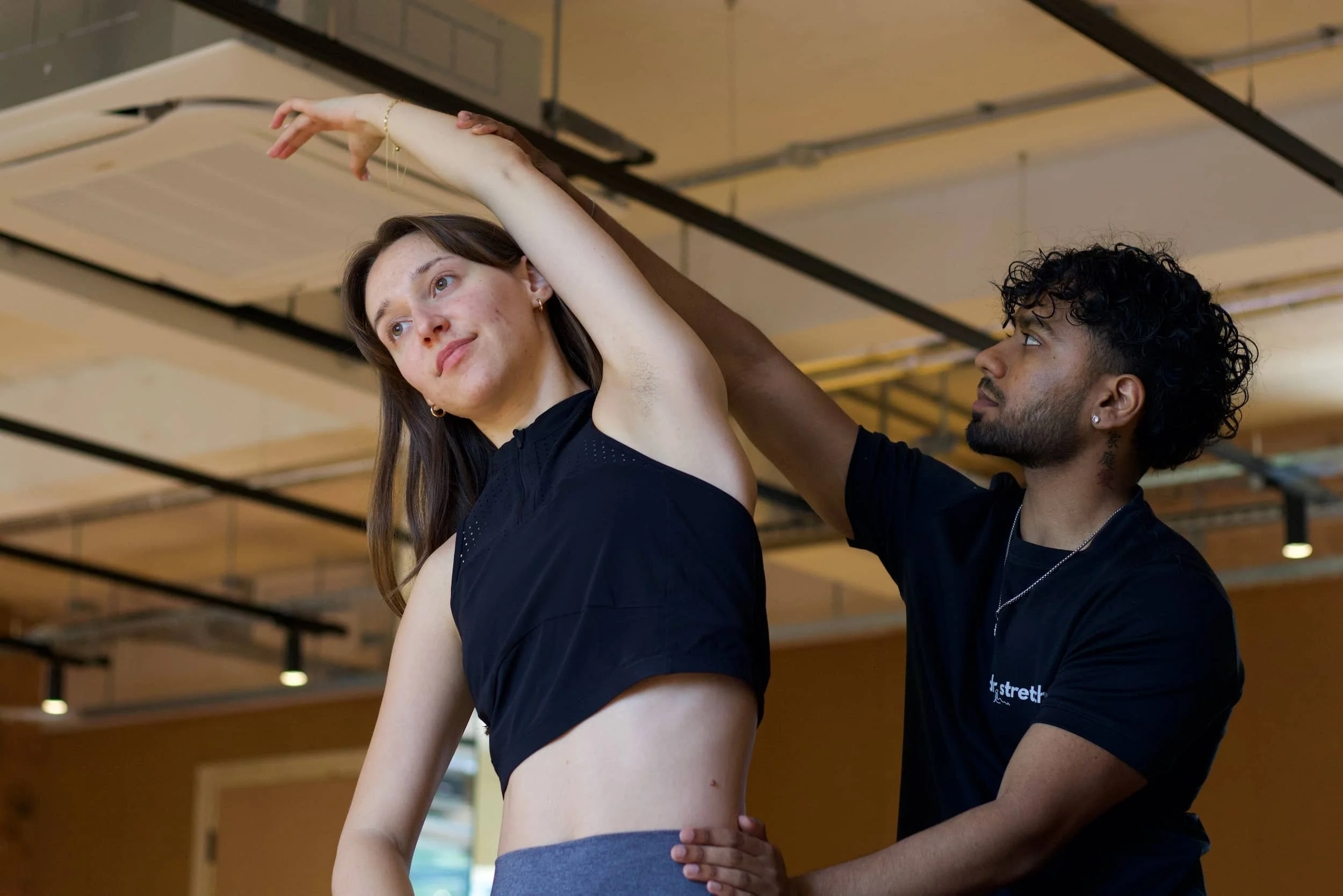 A woman stretching with a trainer assisting her in an indoor gym.