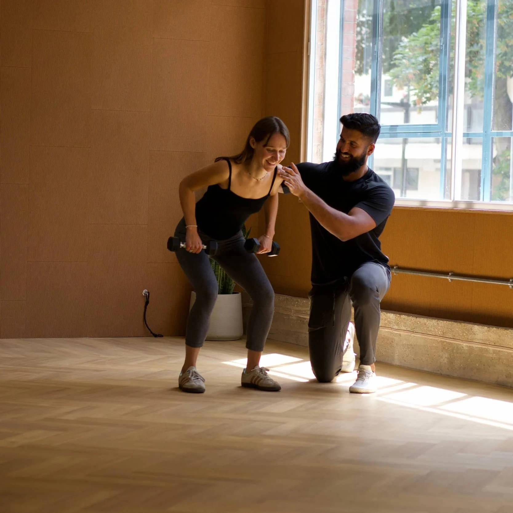 A man assisting a woman with a dumbbell exercise in a bright room with wooden floors and wall, large windows, and natural daylight.