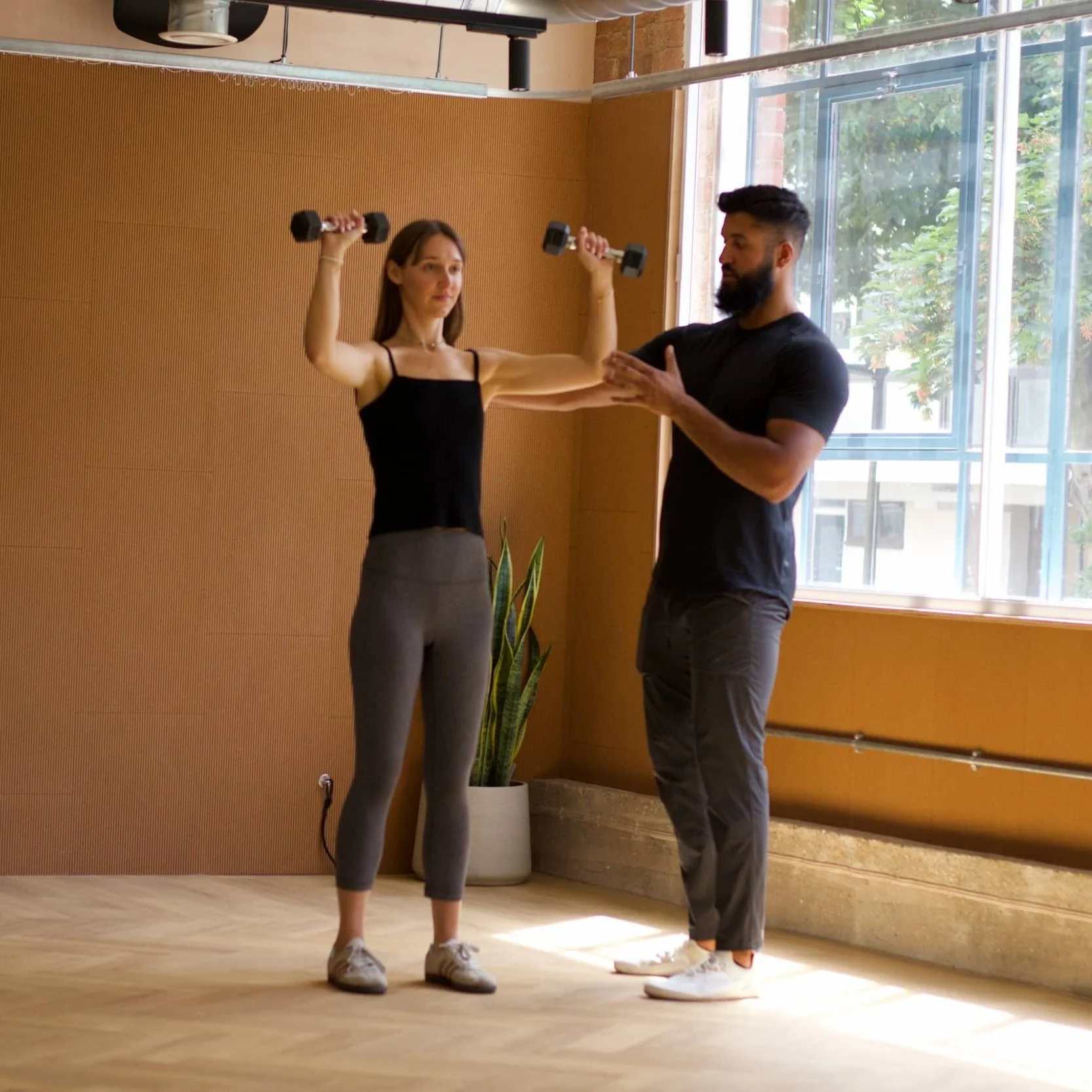 A woman is exercising with dumbbells while a trainer guides her in a room with large windows and wooden walls.