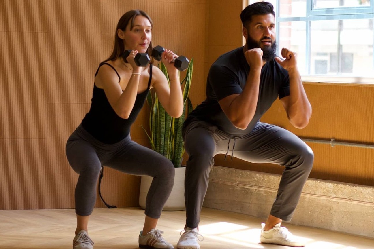 A woman and a man exercising indoors. The woman is holding dumbbells and performing a squat, while the man is also squatting with fists up in a bodybuilding pose. They are in a room with wooden walls, a large window, and a potted plant.
