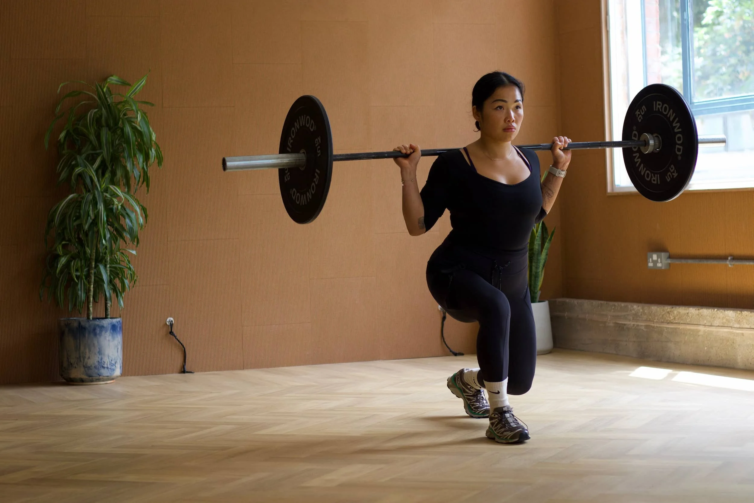 A woman in athletic clothing performing a squat with a barbell in a bright indoor gym or studio.