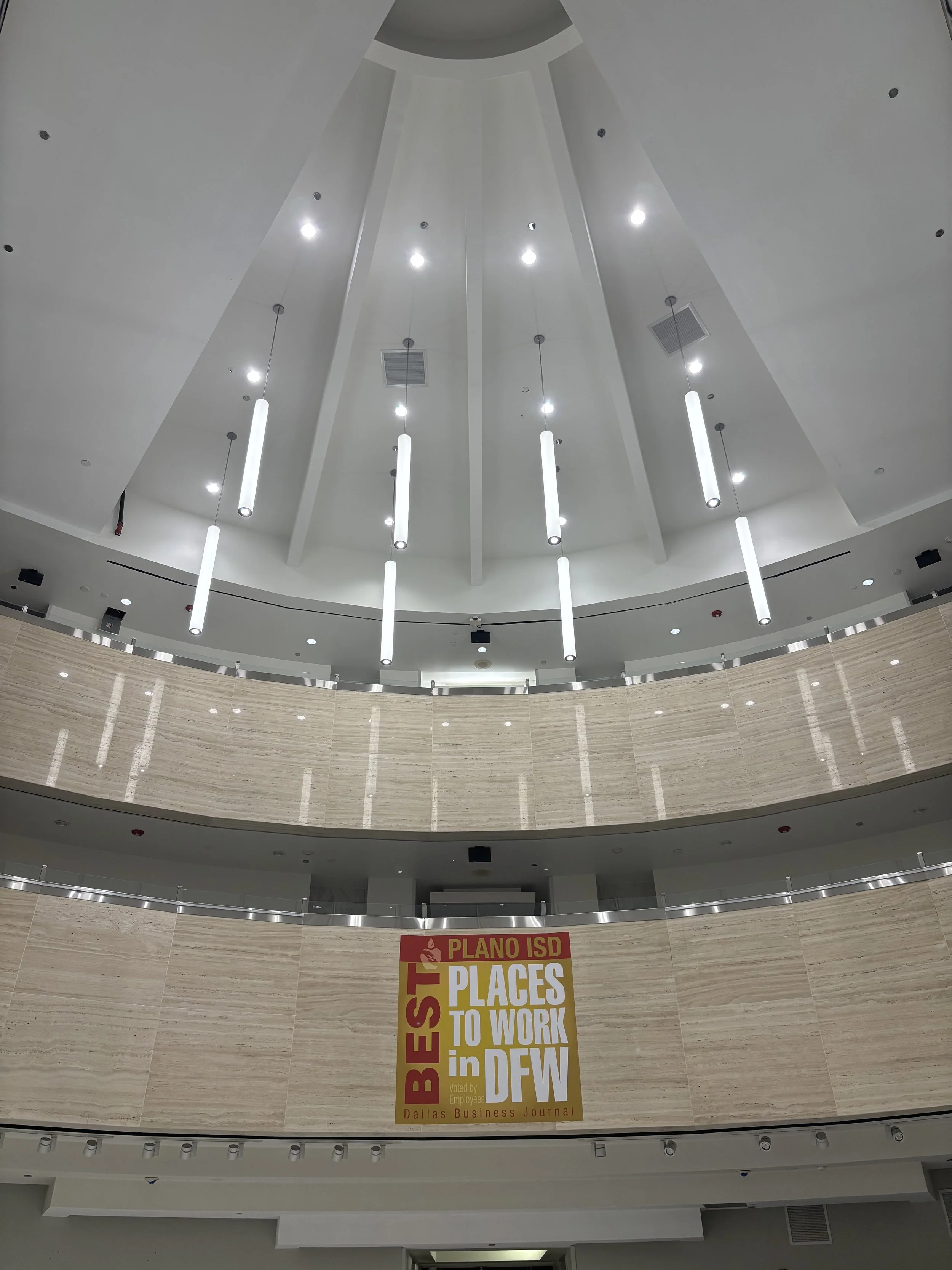 Modern lobby with a high white ceiling, suspended cylindrical lights, marble walls, and a Dallas Business Journal “Best Places to Work in DFW” award sign.