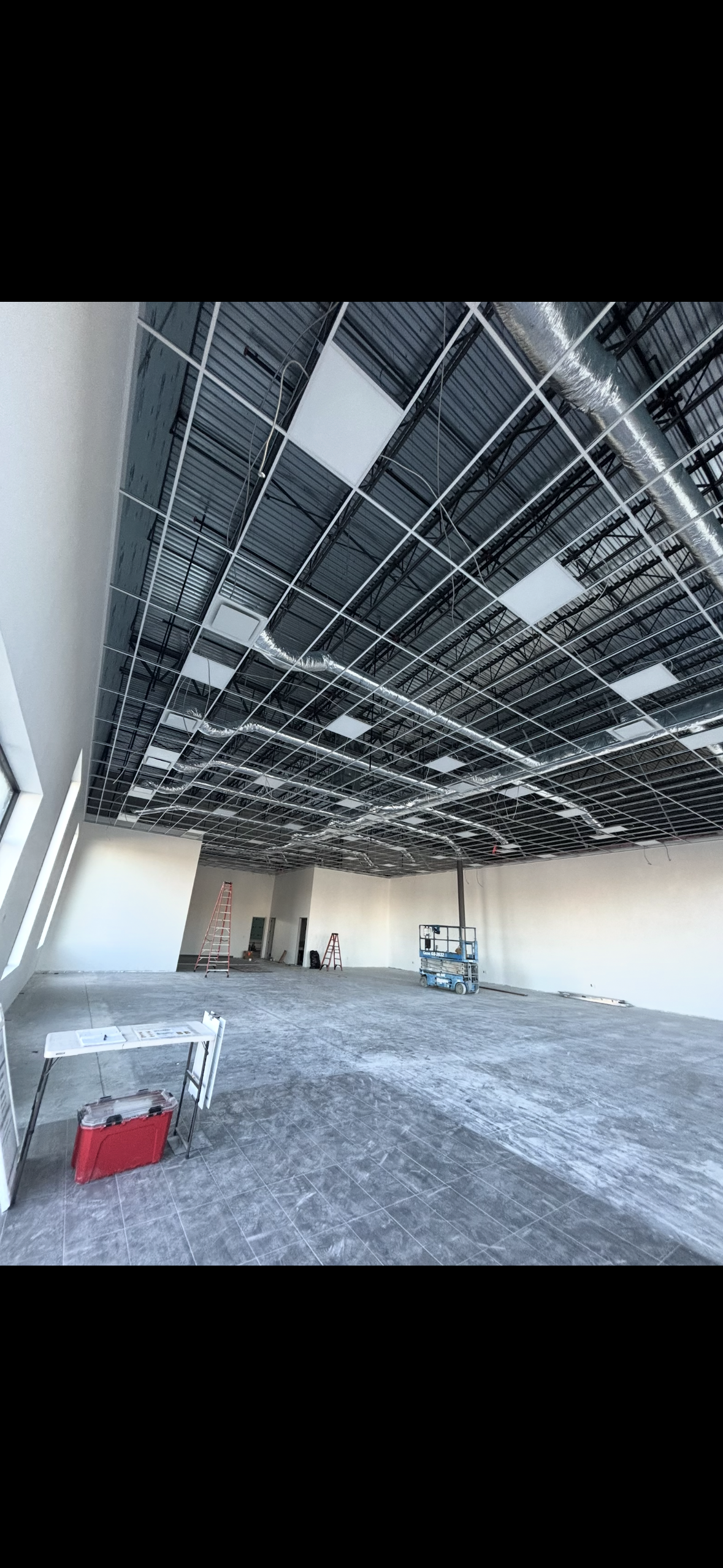 Commercial interior under construction with exposed ceiling grid, HVAC ductwork, lighting panels, ladders, and a scissor lift