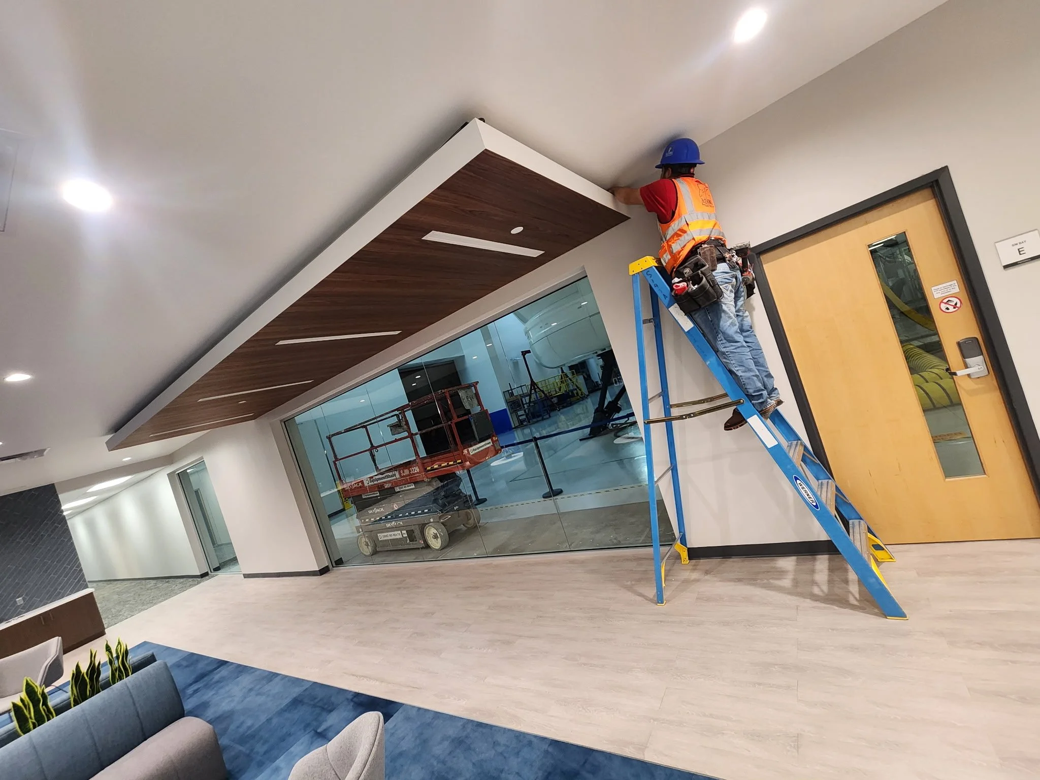 Construction worker in a safety vest standing on a ladder while installing ceiling trim in a modern office lobby