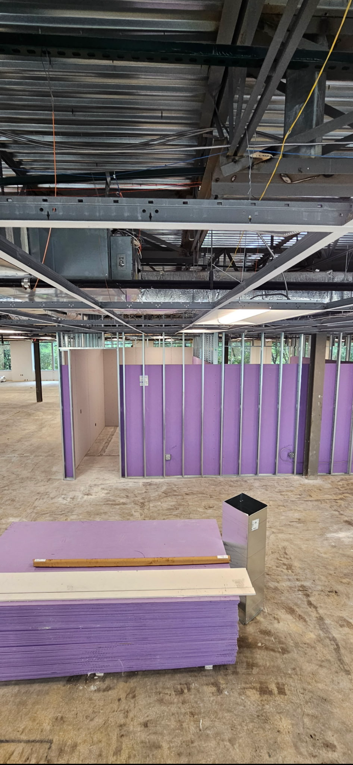 Commercial building interior under construction with exposed ceiling, wiring, metal framing, purple drywall partitions, and stacked drywall sheets