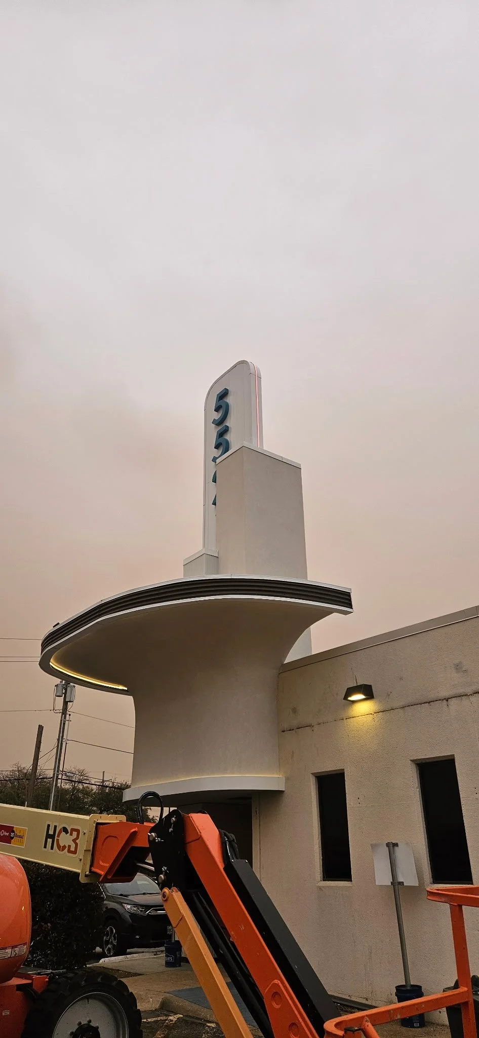 Art Deco-style building entrance with a curved canopy and vertical "5" sign, viewed at dusk behind orange construction equipment