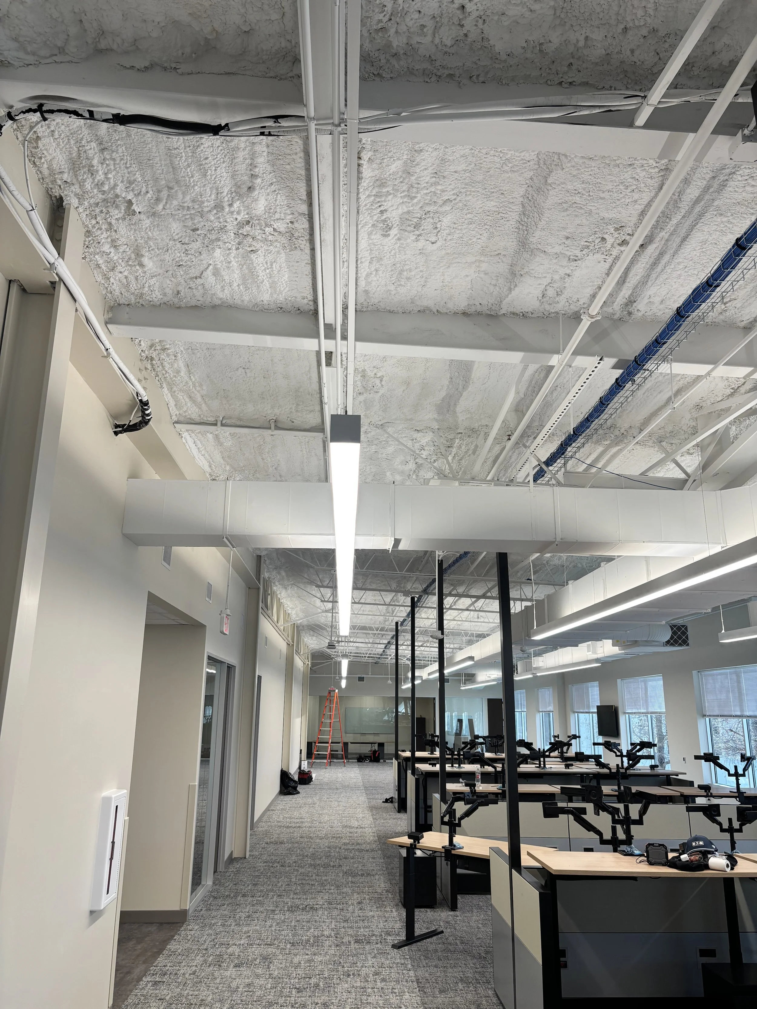 Modern open-plan office with rows of desks, exposed spray-foam ceiling, ductwork, cable trays, pendant lighting, windows, and an orange ladder.