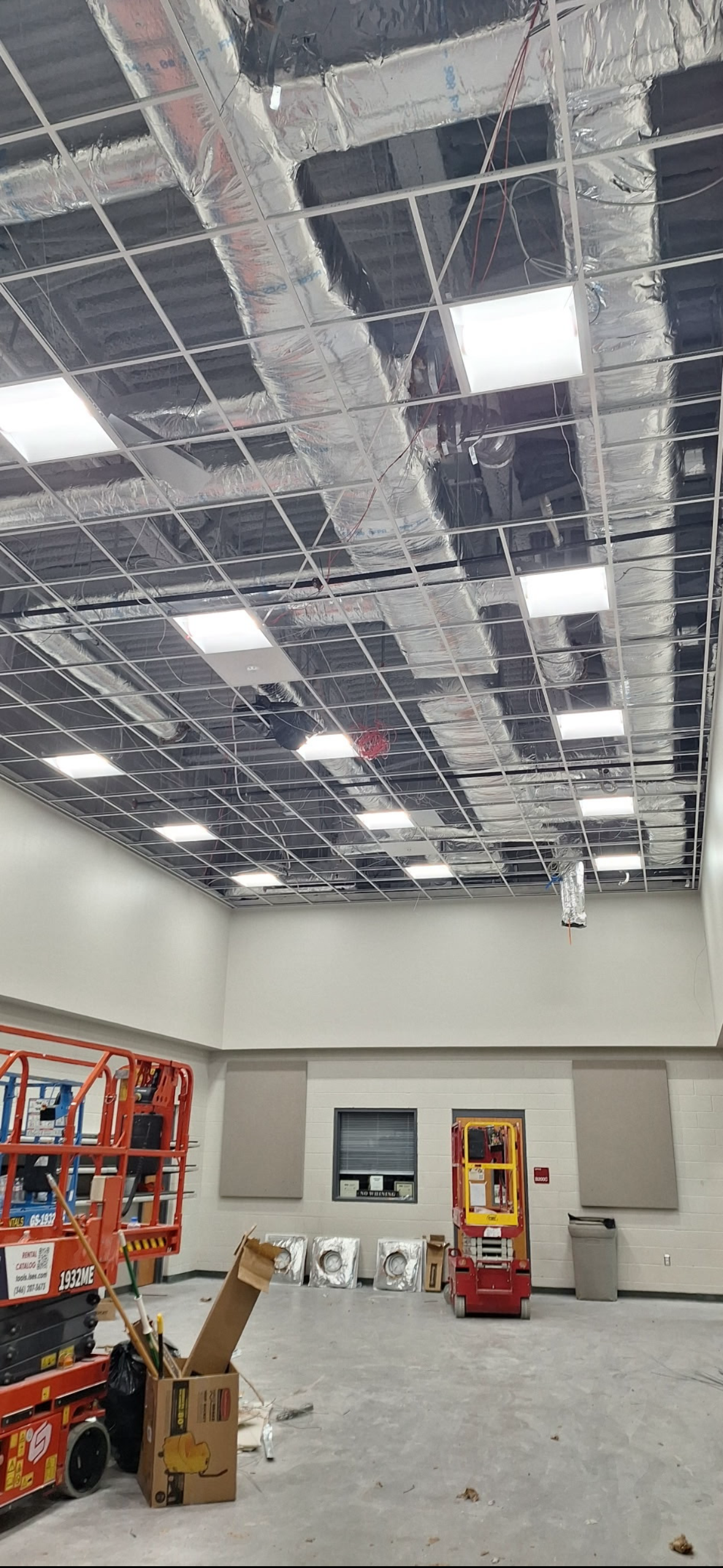 Commercial room under construction with exposed HVAC ducts, suspended ceiling grid, fluorescent lights, scissor lifts, and construction materials