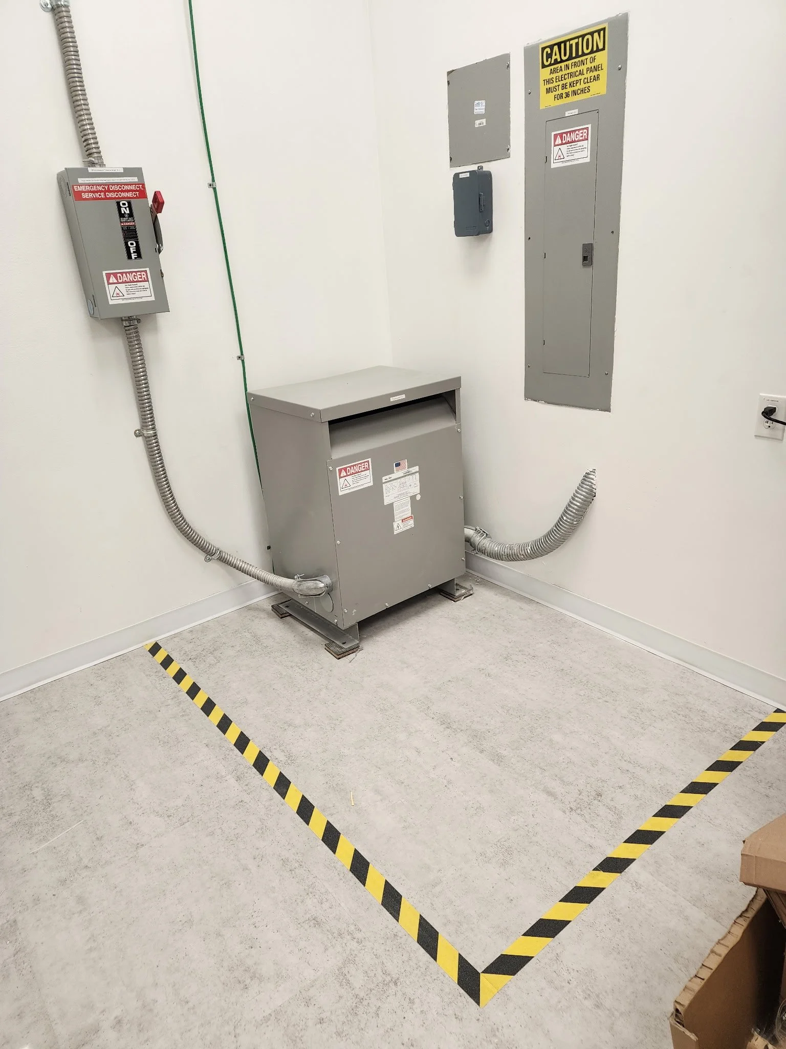 Industrial electrical equipment in a utility room, including a disconnect switch, control cabinet, warning signs, conduit, and a black-and-yellow safety boundary.