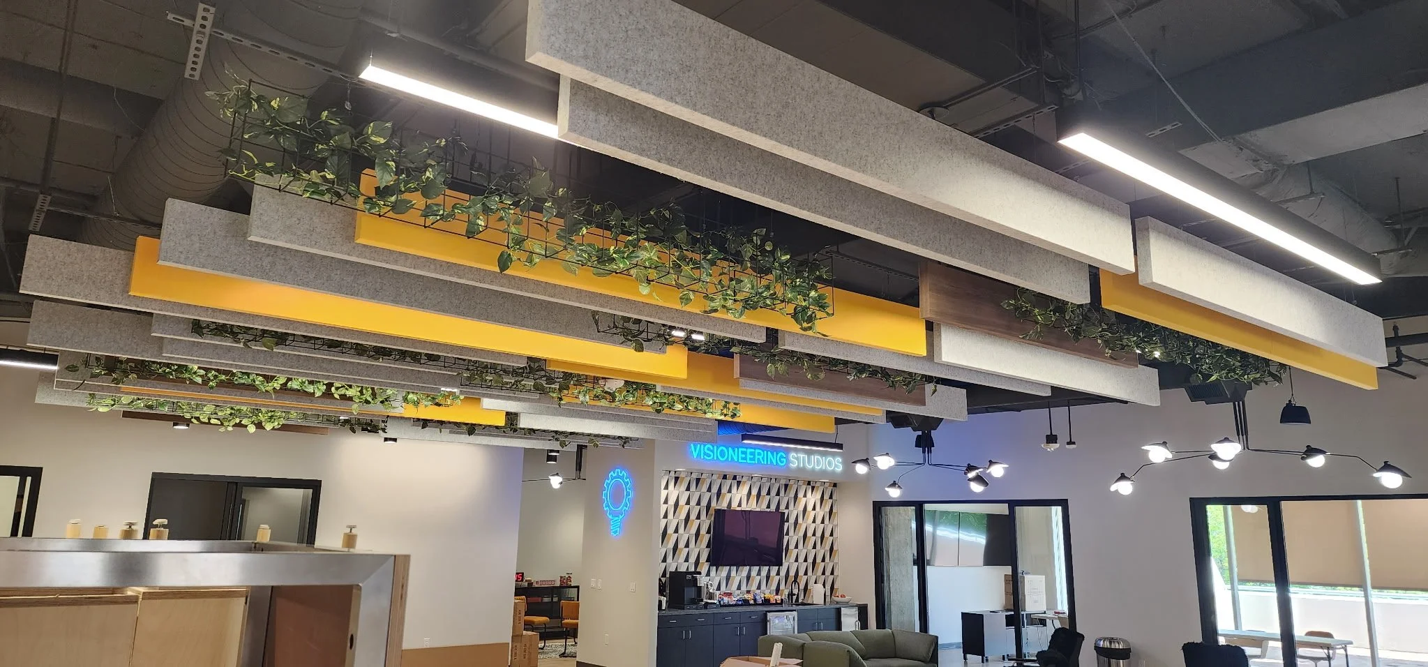 Modern office lounge with yellow and gray suspended ceiling panels, hanging greenery, pendant lighting, kitchenette, and Visioneering Studios sign