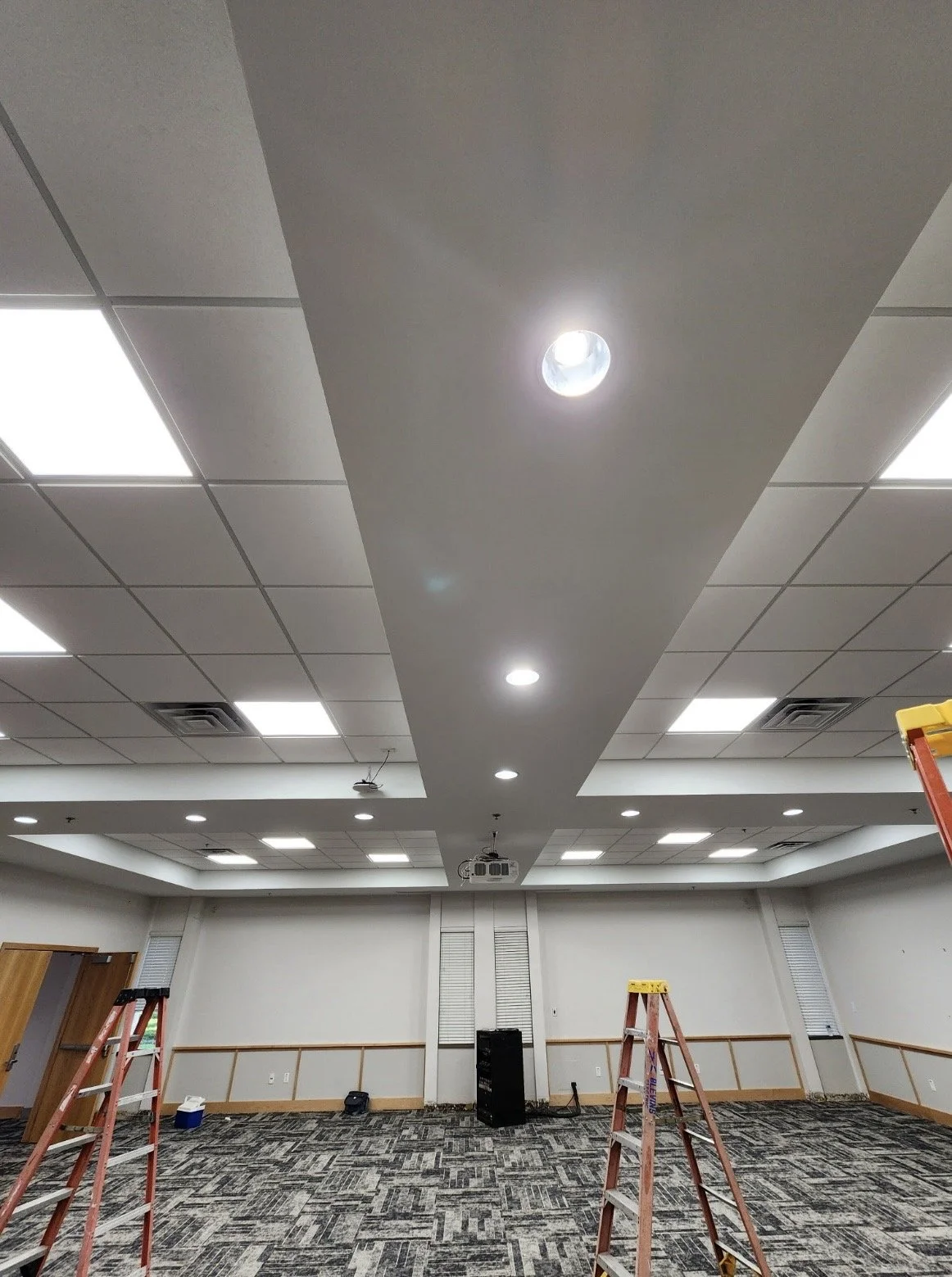 Empty commercial room with drop ceiling lights, recessed lighting, carpeted floor, and several ladders