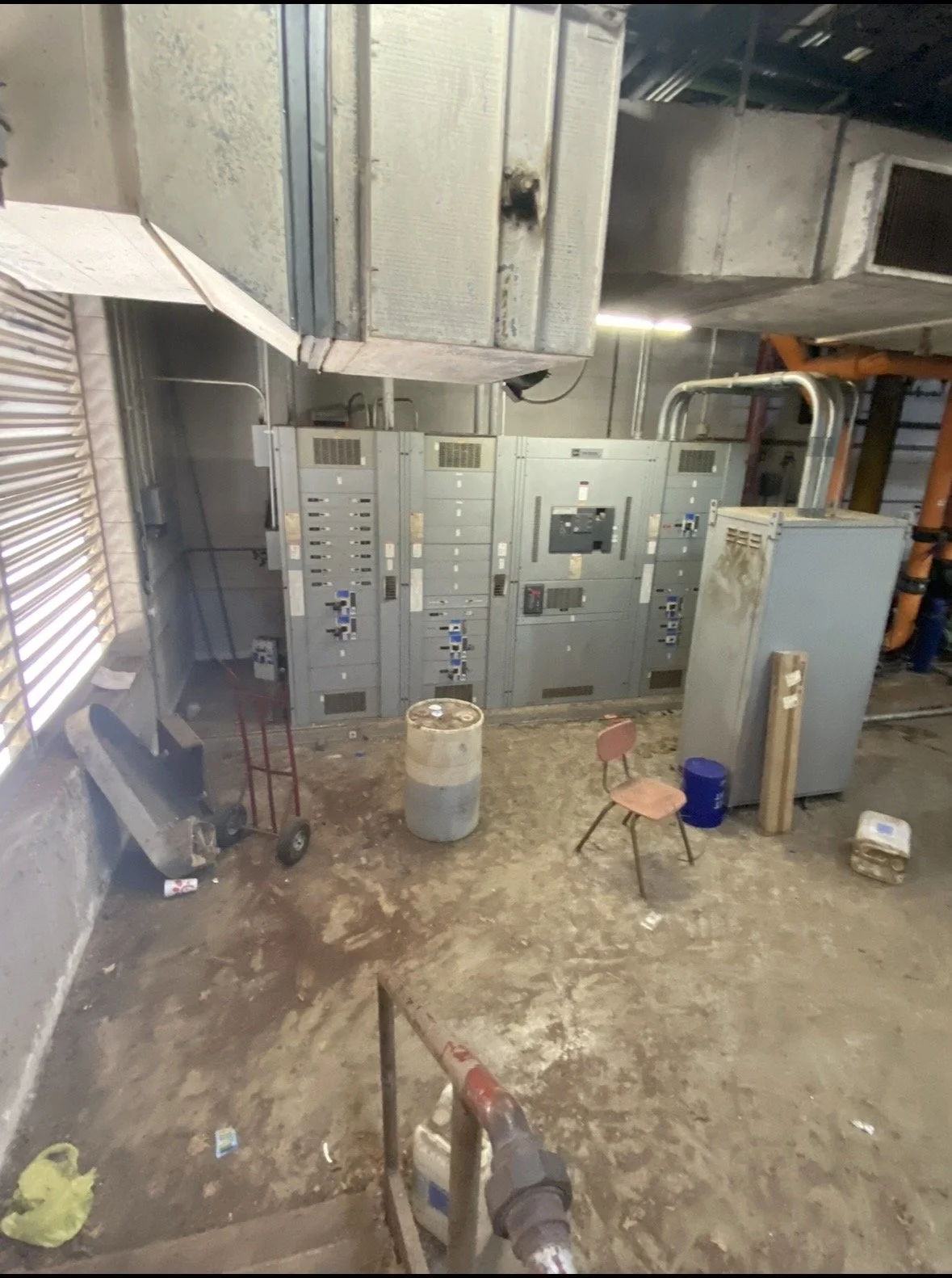 Industrial electrical switchgear panels in a utility room with overhead ductwork, pipes, a barrel, chair, and hand truck